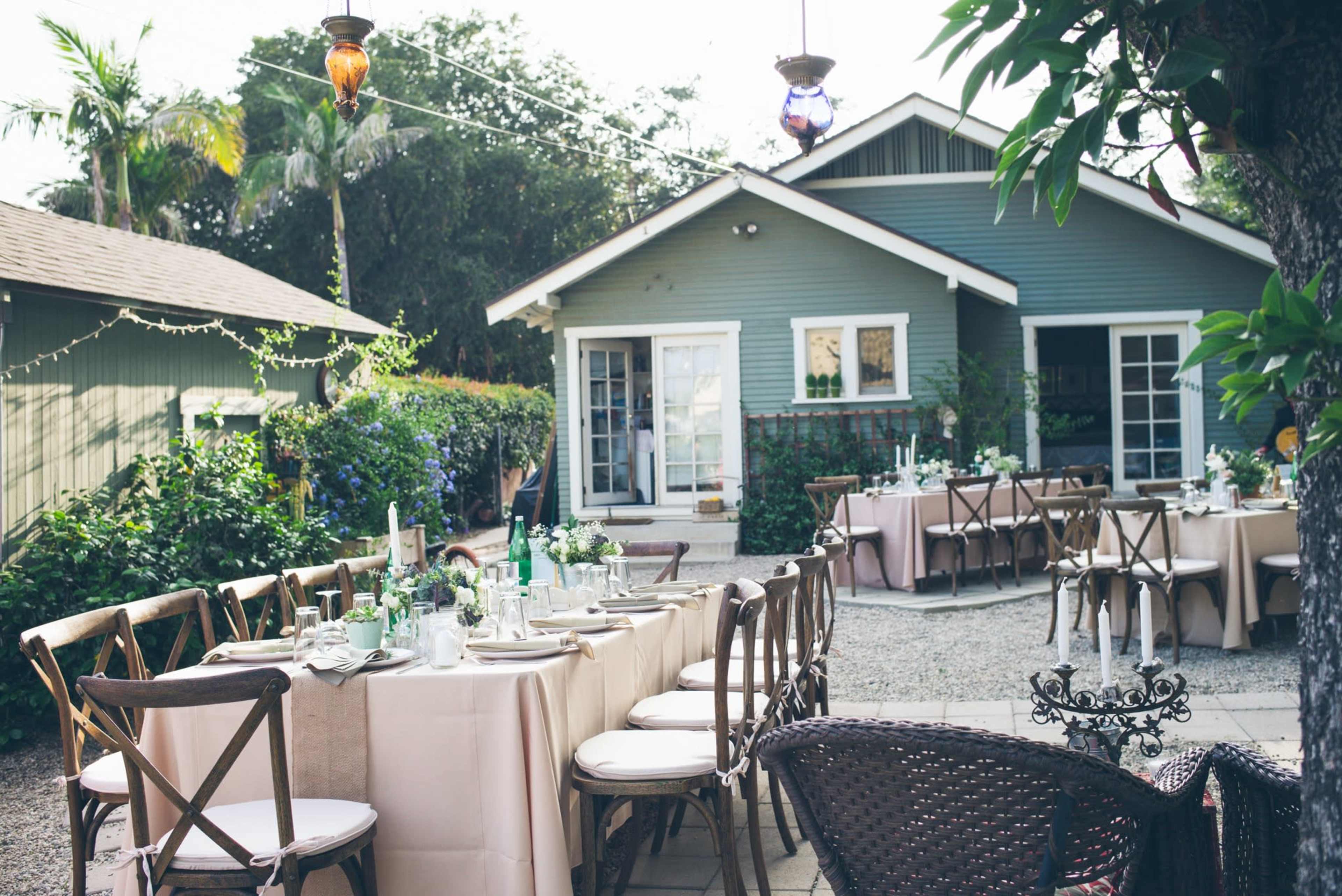 The image shows an outdoor dining setup with elegantly arranged tables and chairs in front of a green house surrounded by trees and decorative lights.