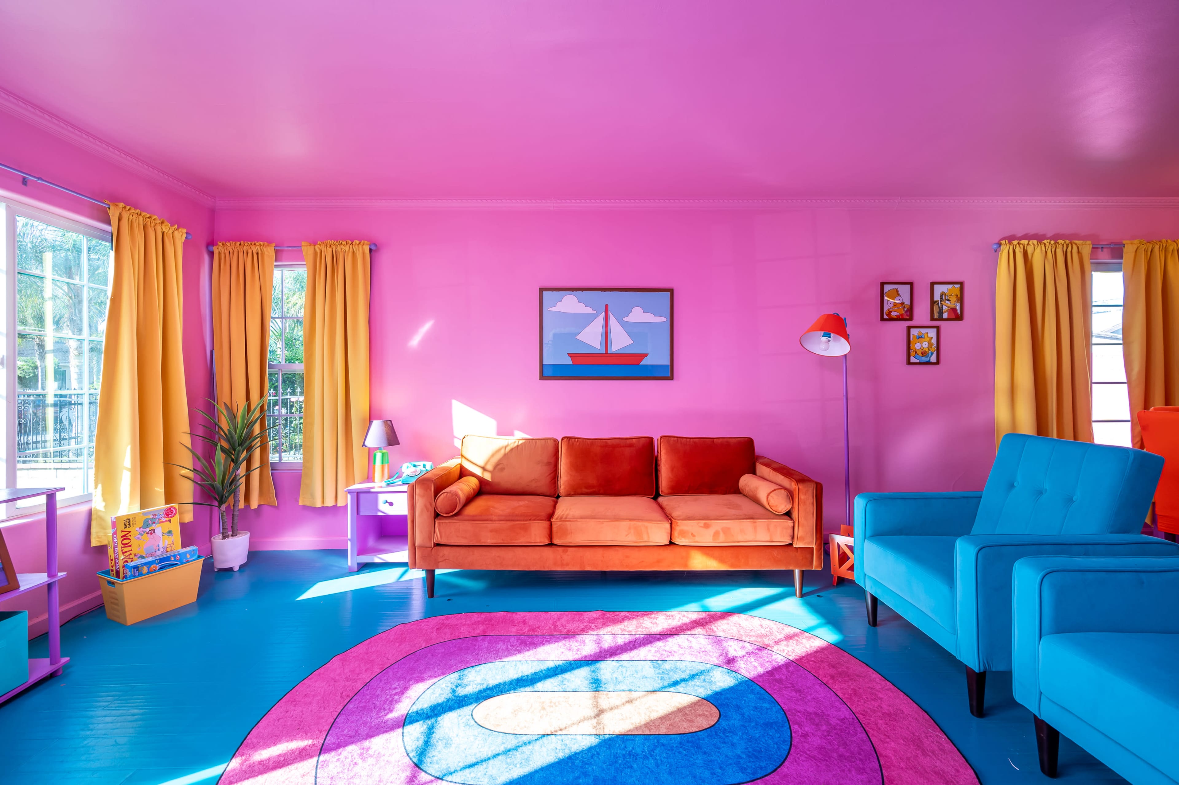 The scene features a brightly colored living room with pink walls, an orange sofa, blue chairs, and a circular rug, illuminated by natural light from large windows.