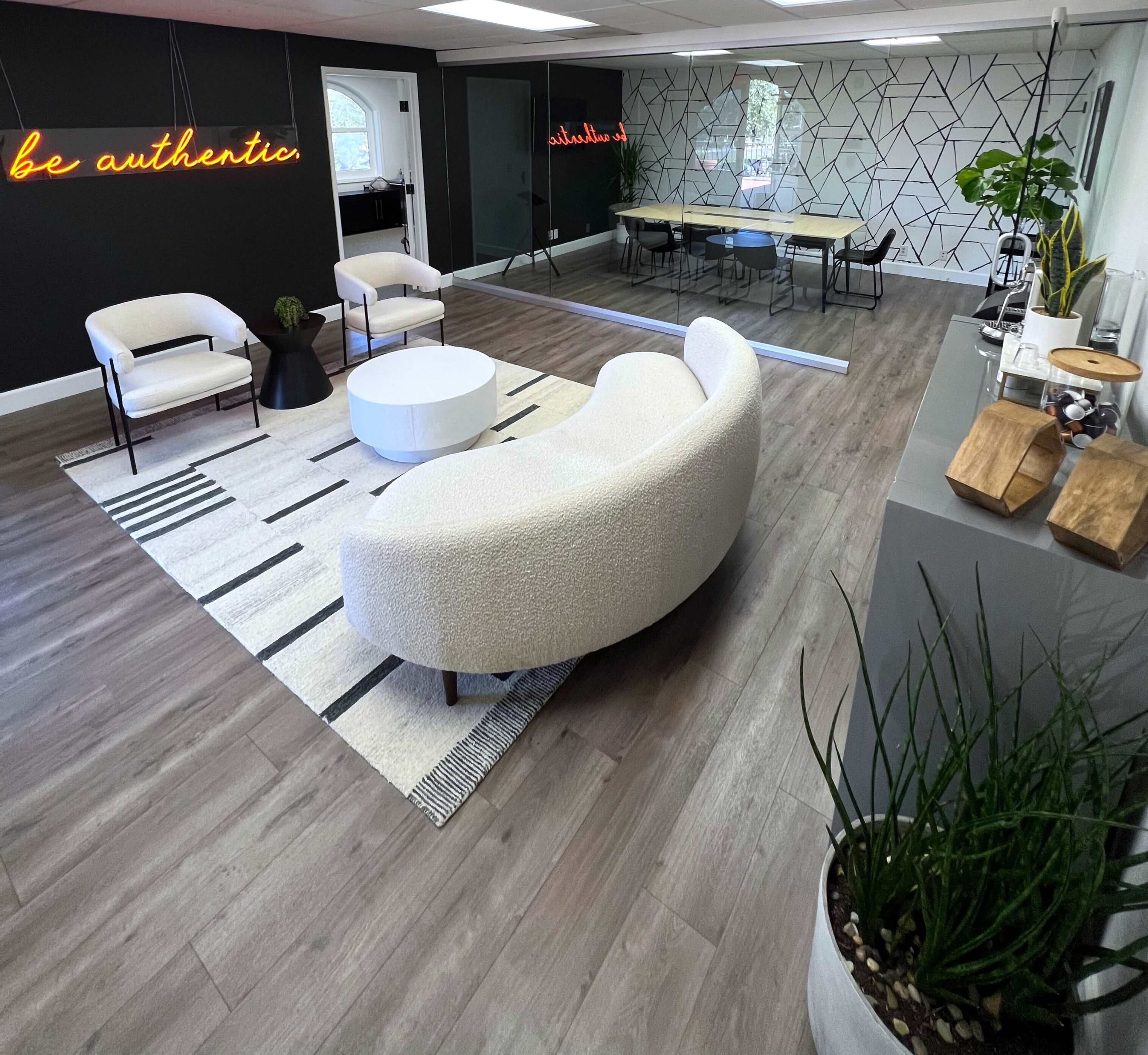 A modern office space with a curved sofa, two chairs, a round coffee table, and a meeting area featuring a large table against a geometric-patterned wall.
