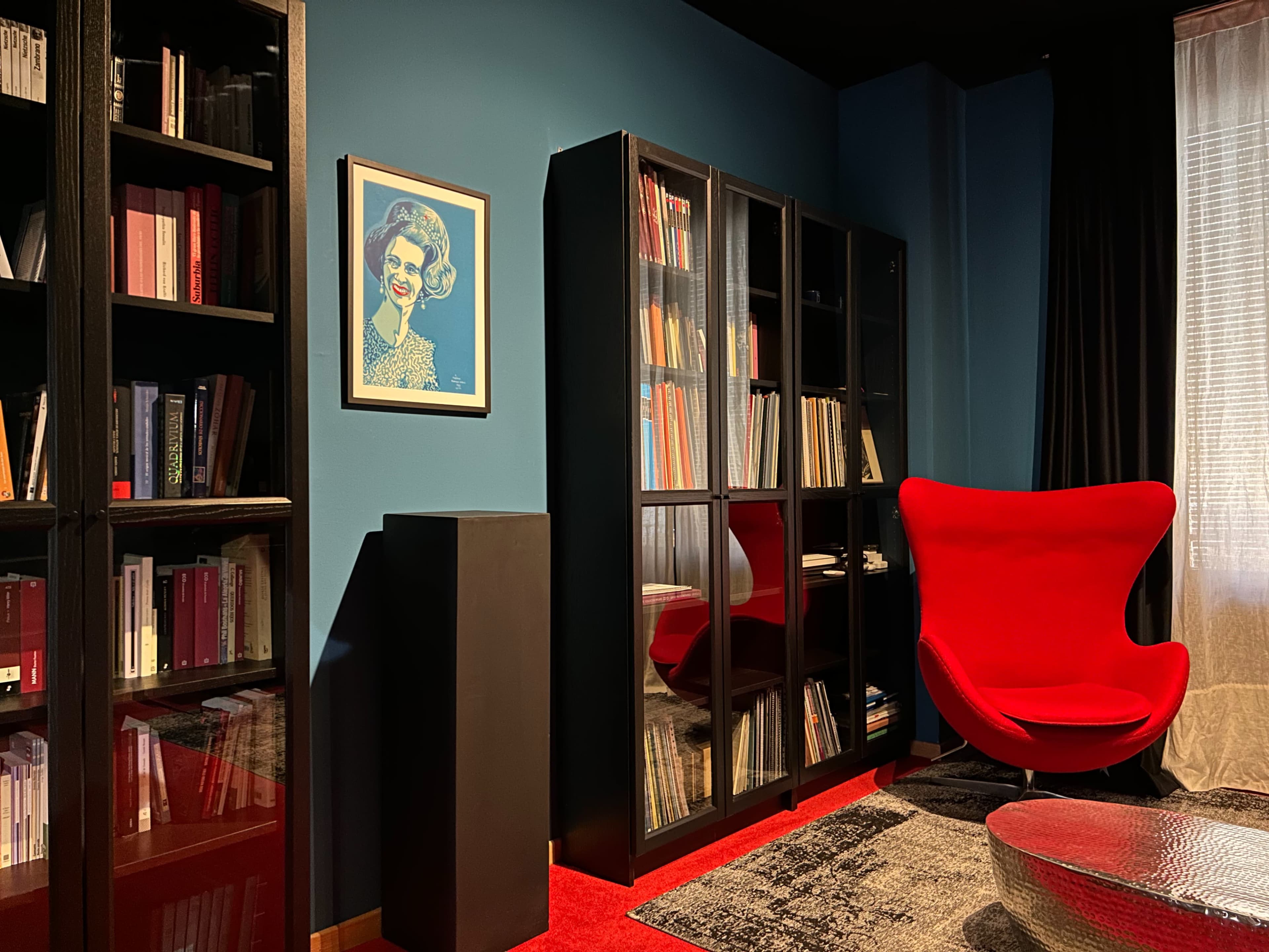 The image shows a modern room with dark blue walls, a red chair, a glass-fronted bookshelf filled with books, and a framed artwork on the wall.