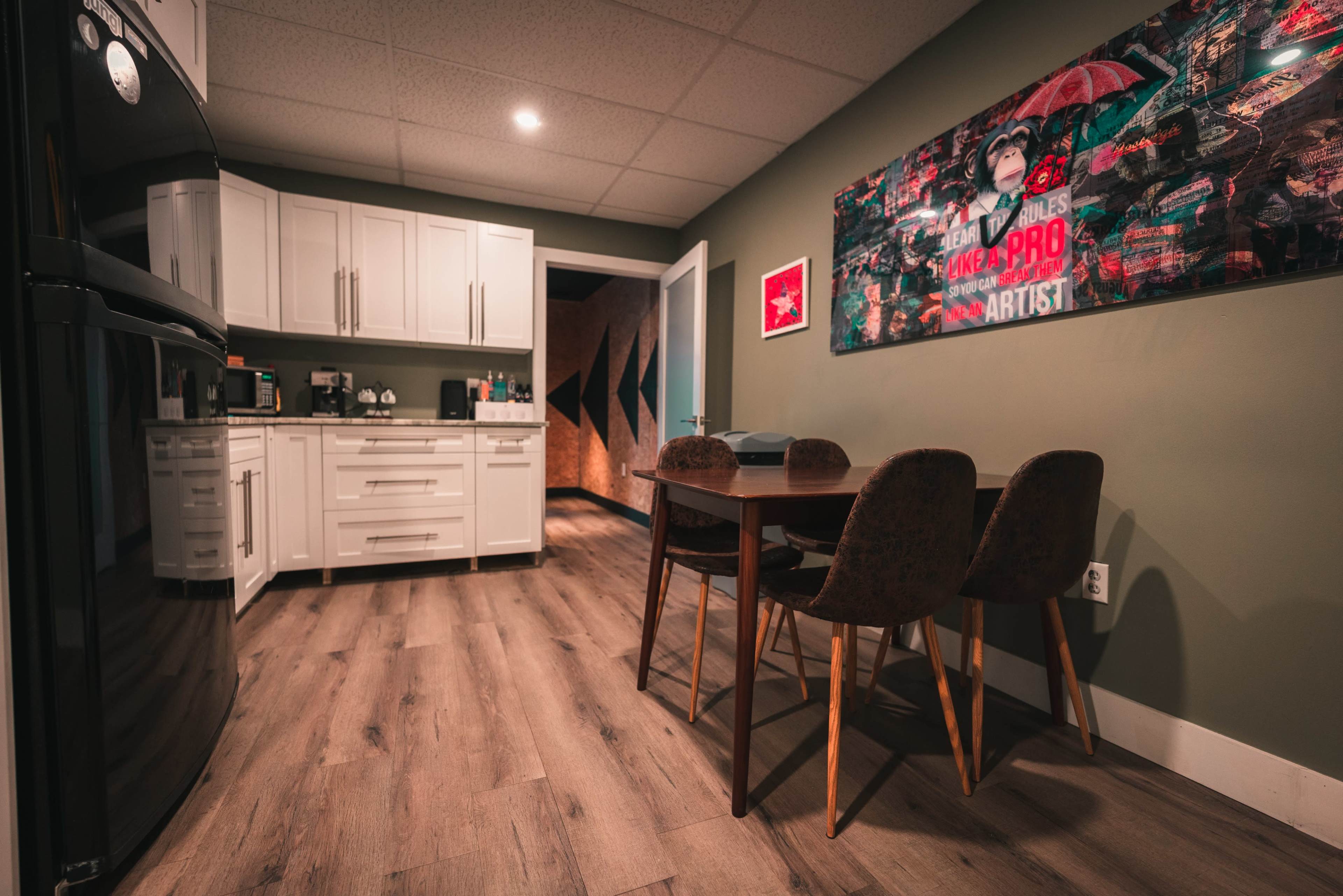 A modern kitchen area features white cabinets, a black refrigerator, a dining table with four chairs, and a vibrant artwork on the wall.