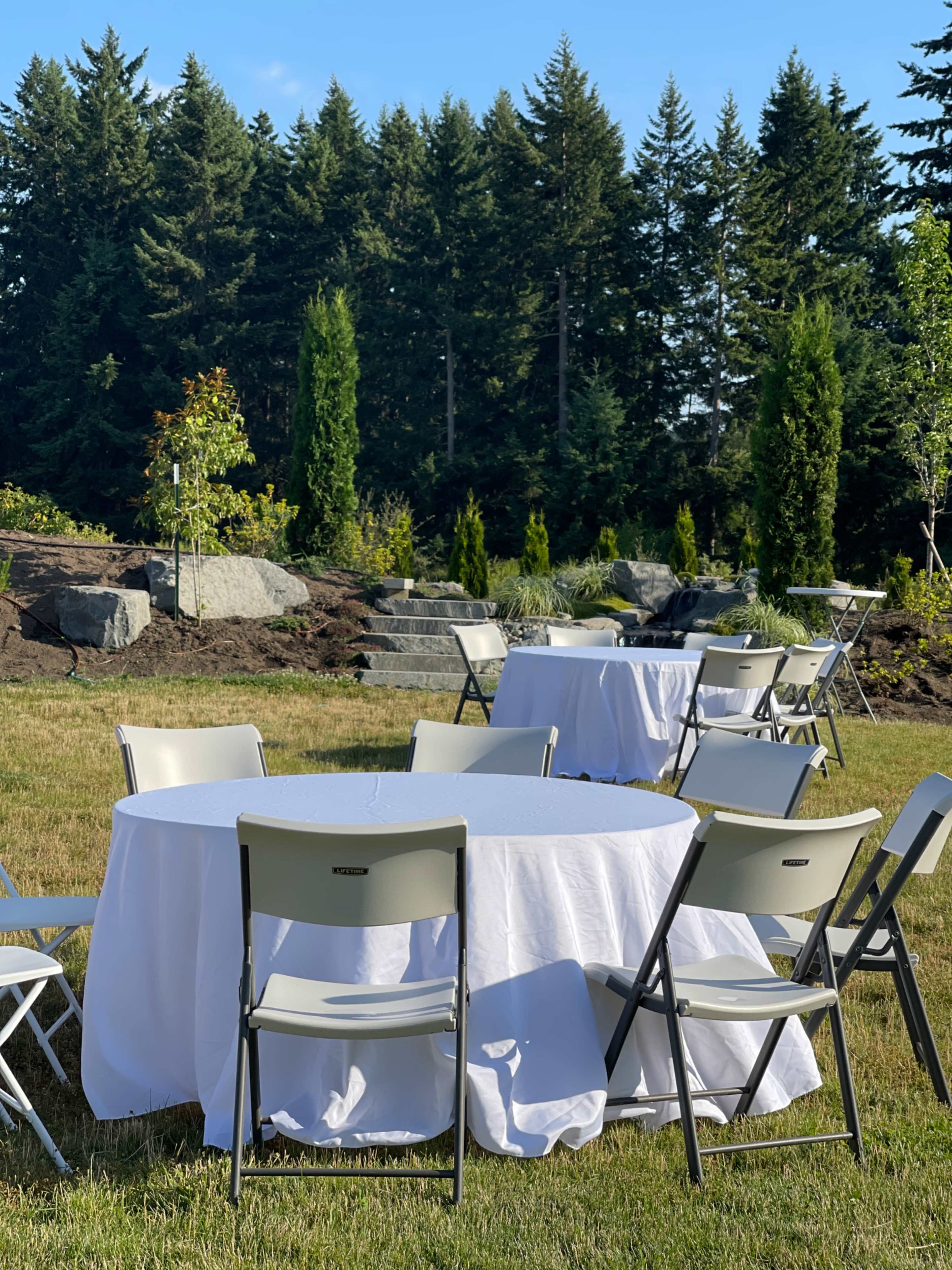 Tables covered with white tablecloths and surrounded by folding chairs are arranged on a grassy area with trees in the background.