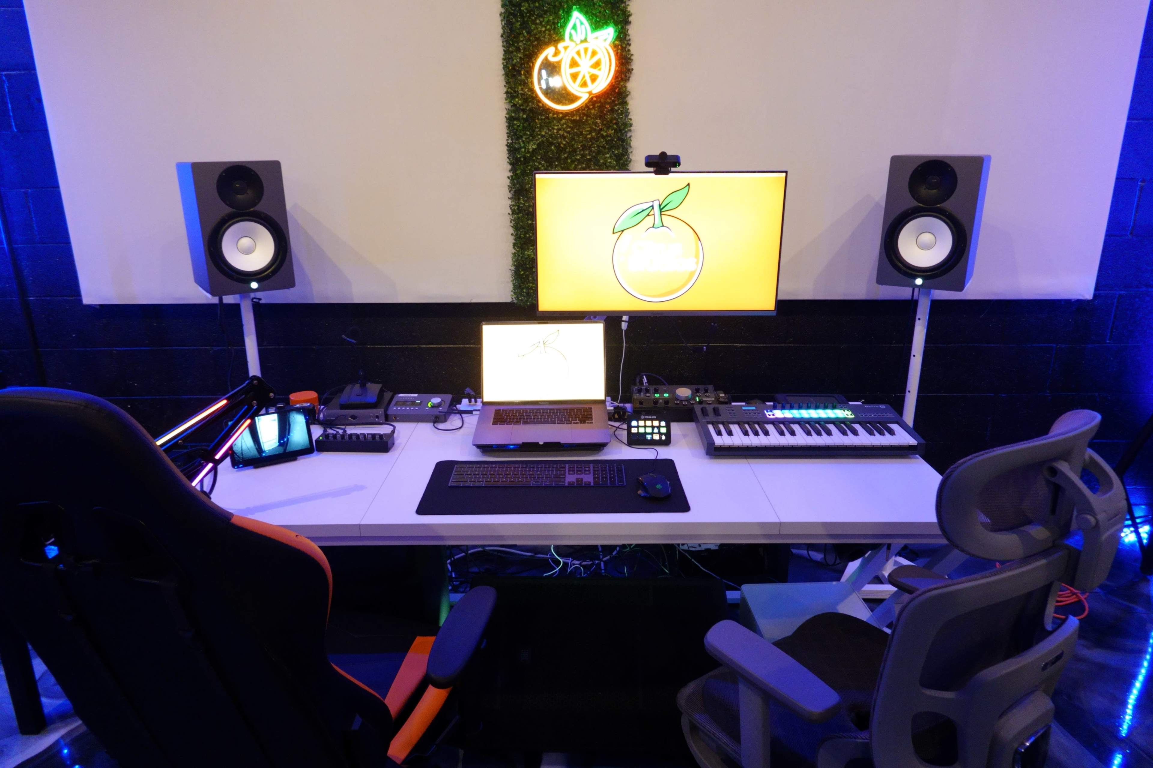 The image shows a modern home studio setup featuring a desk with a computer monitor, laptop, MIDI keyboard, and speakers, illuminated by an orange-themed neon sign.
