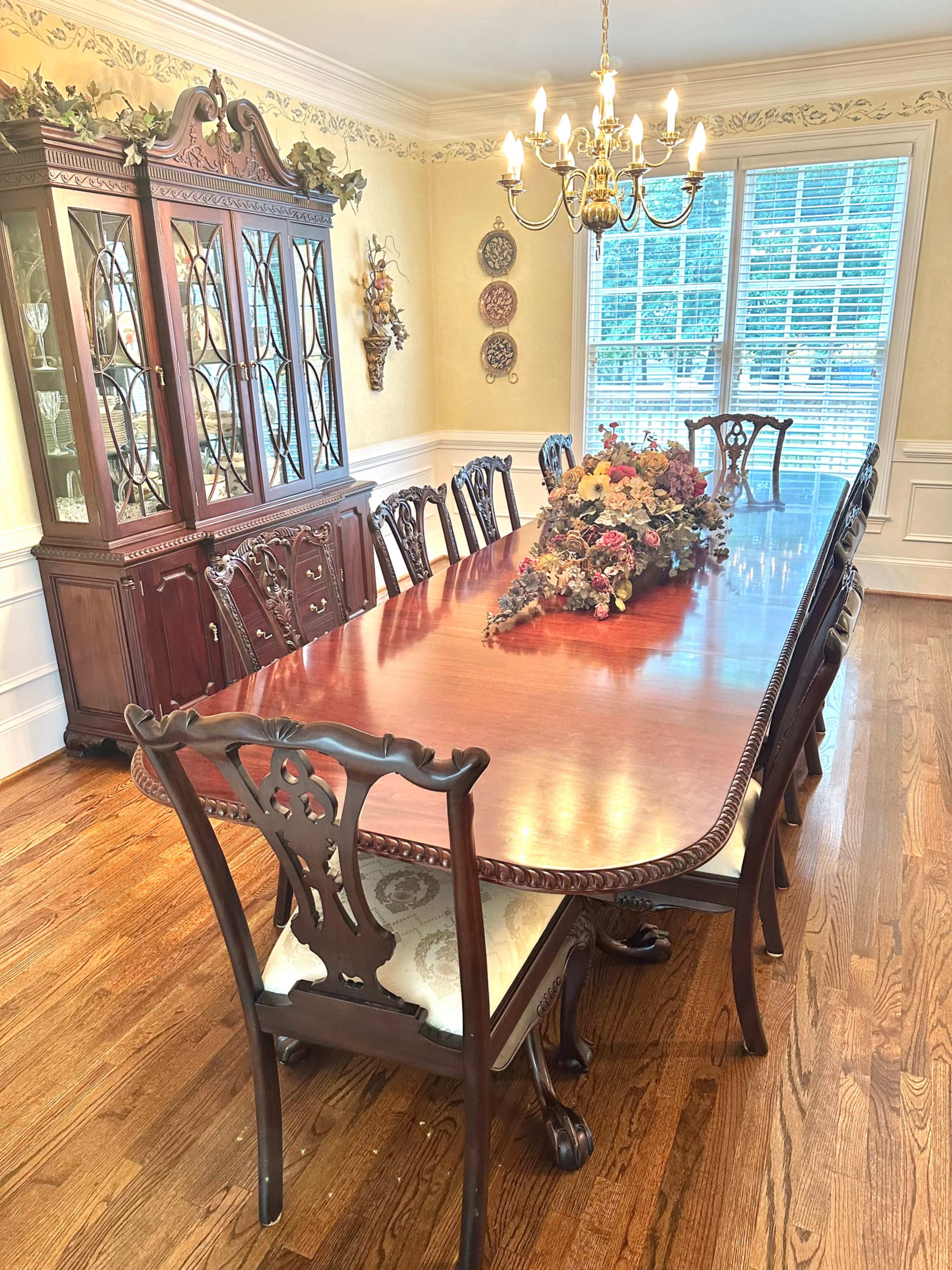 A large wooden dining table surrounded by intricately carved chairs is set in a well-lit dining room, accompanied by a china cabinet and decorative accents on the wall.