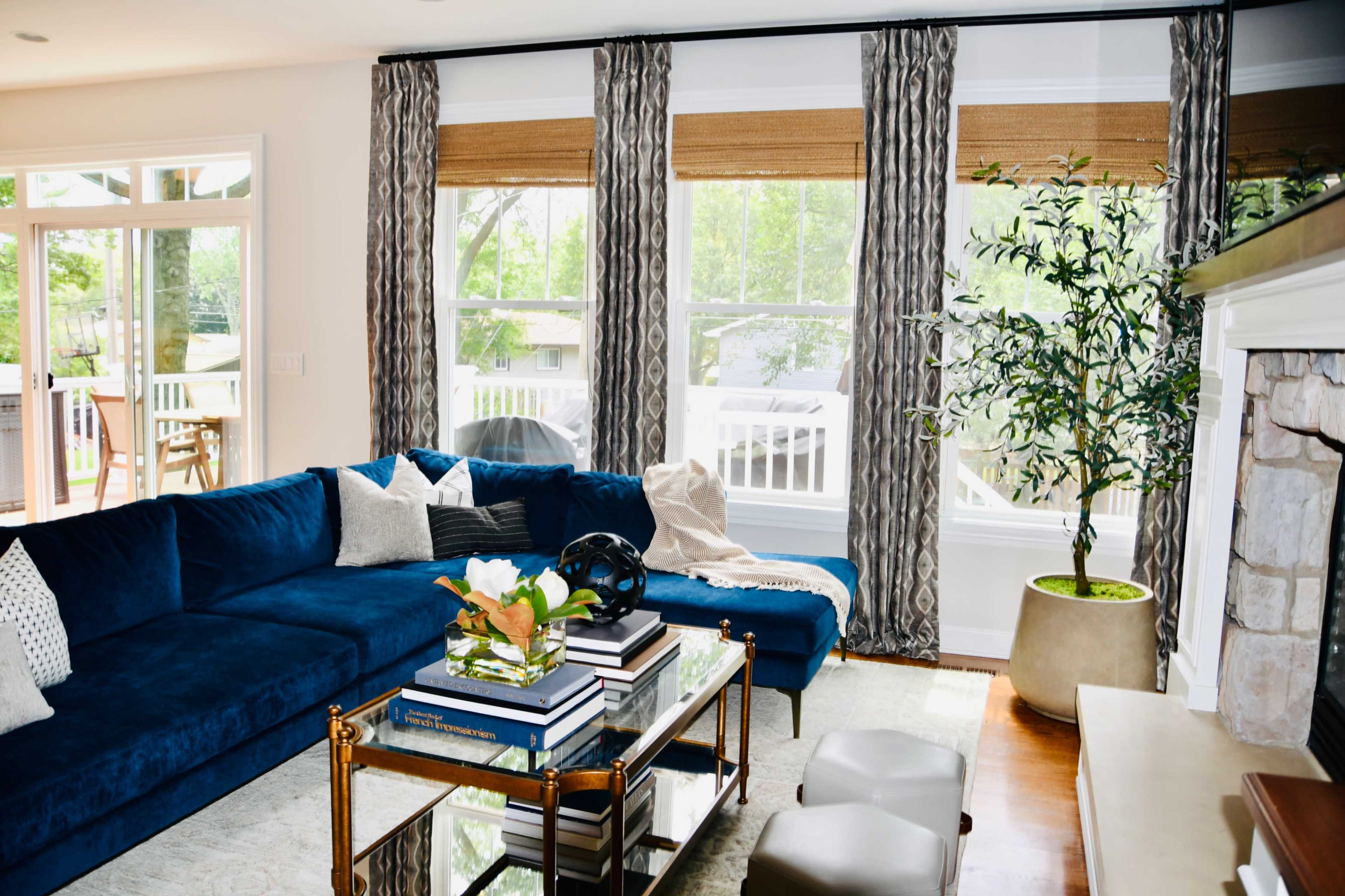 A blue sectional sofa is positioned in a bright living room with large windows, adorned with decorative pillows and a coffee table stacked with books and a plant.