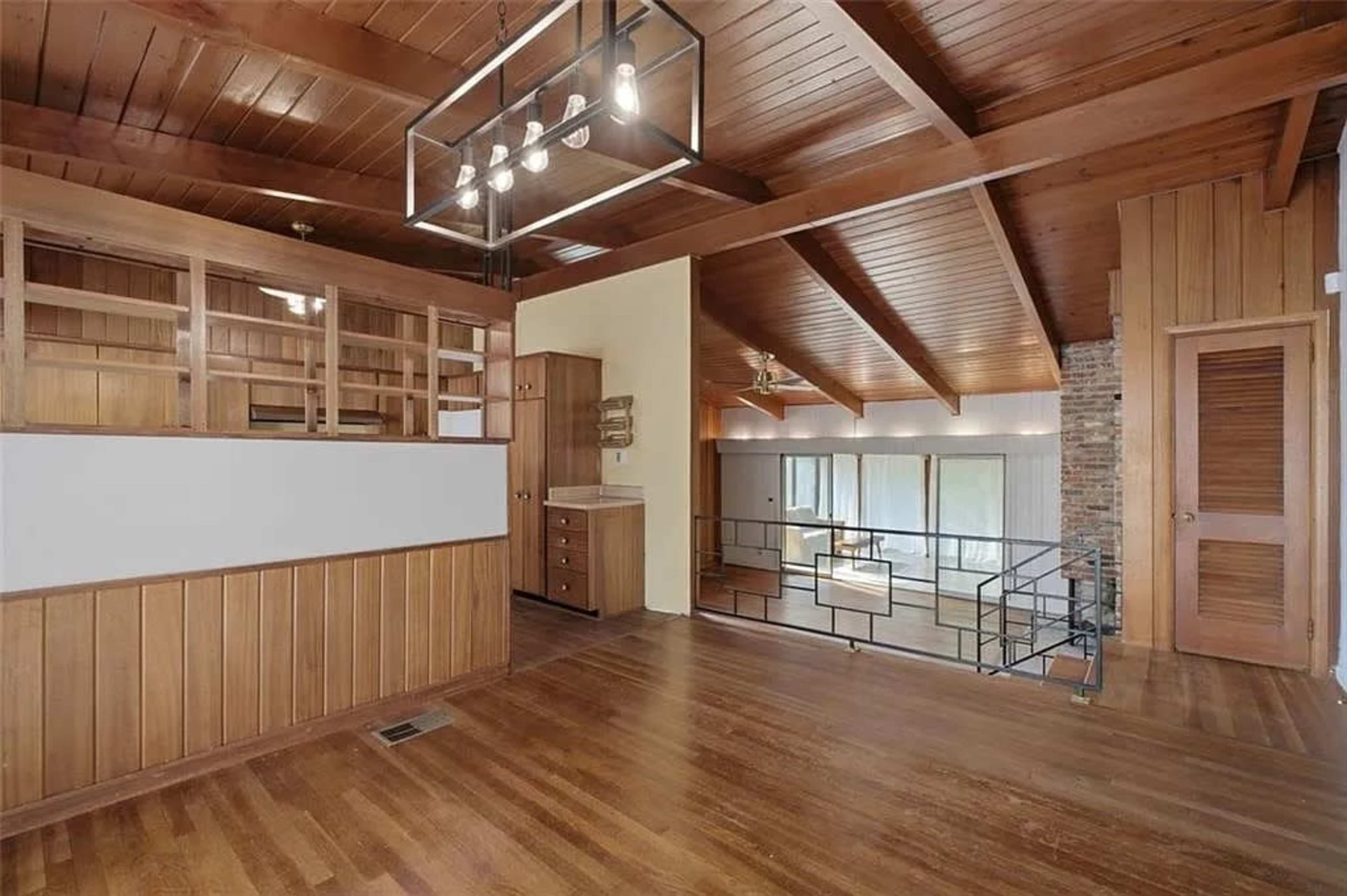 The image shows a spacious room with wooden flooring, exposed beams on the ceiling, and a divided area featuring a kitchen and living space with large windows.