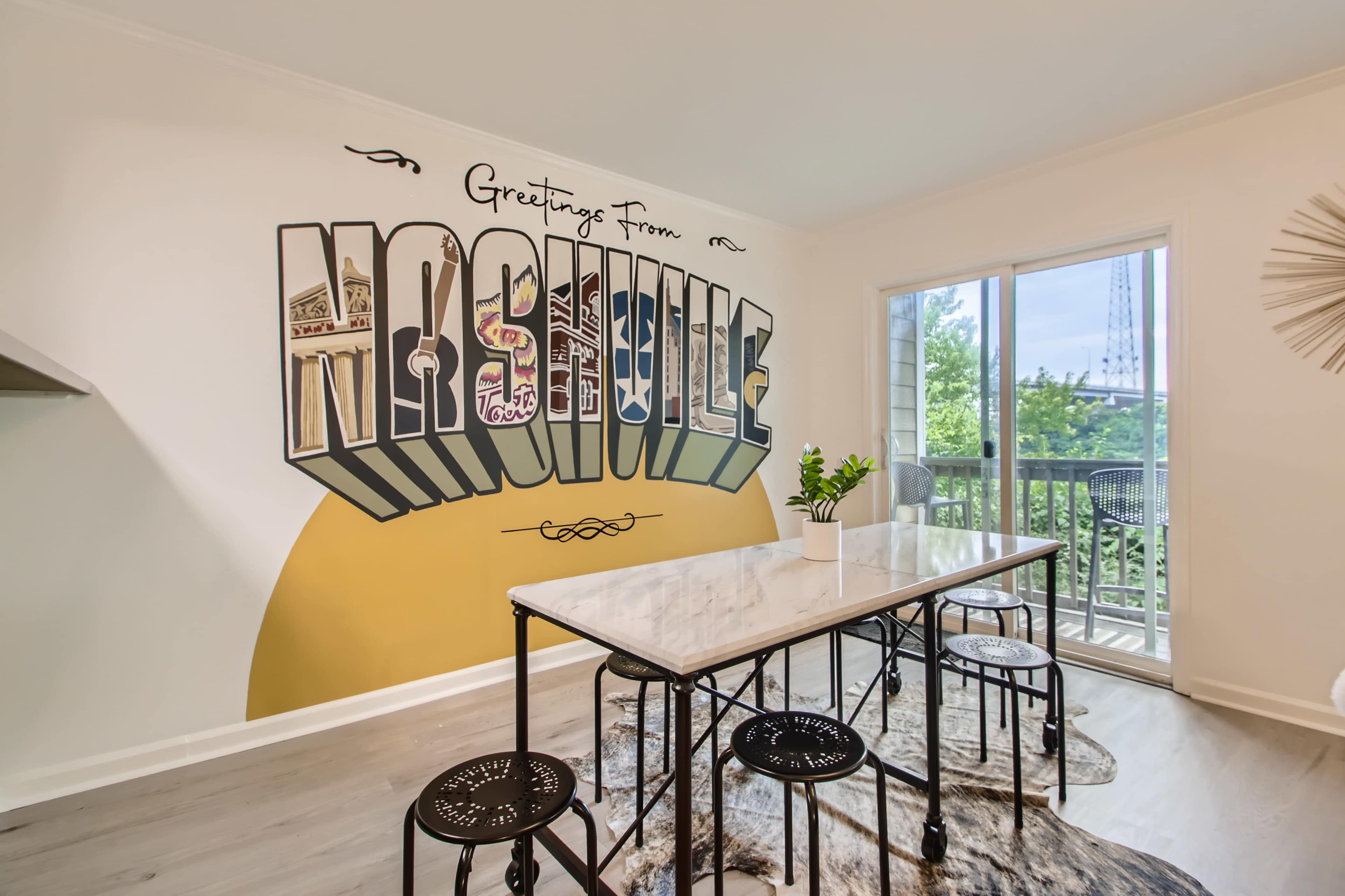 A modern dining area with a large marble table surrounded by black chairs, set against a wall featuring a colorful "Greetings From Nashville" mural and a view of the outdoors through a sliding glass door.