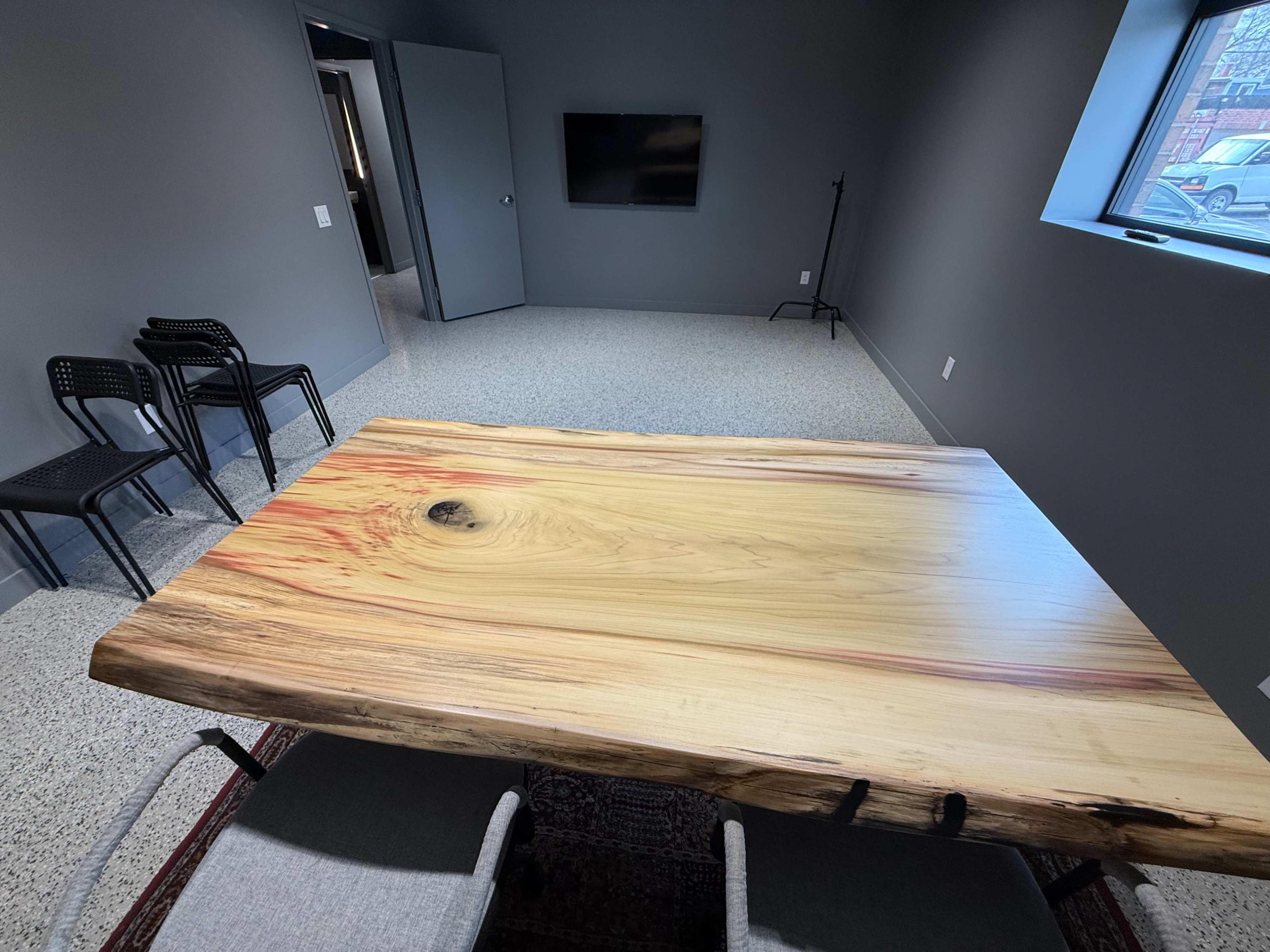 A large, natural wood table with a smooth surface is positioned in a sparsely furnished room featuring a television, a closed door, and several black chairs.