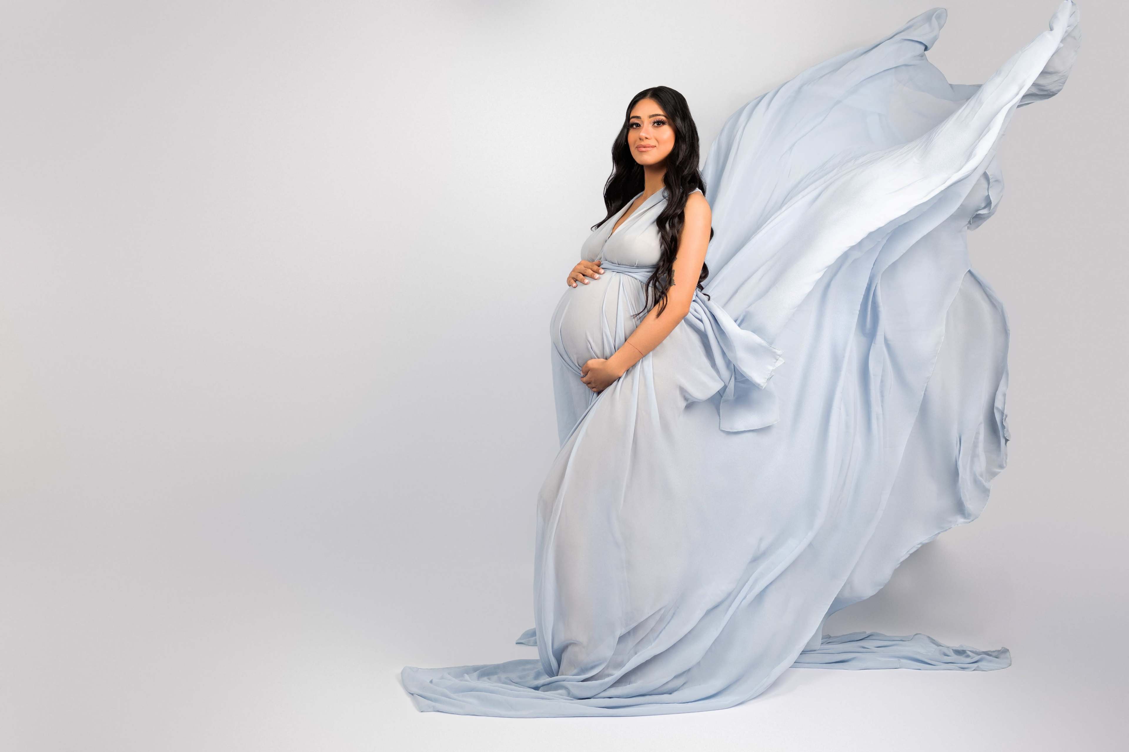 A woman in a flowing blue gown poses gracefully while cradling her pregnant belly.