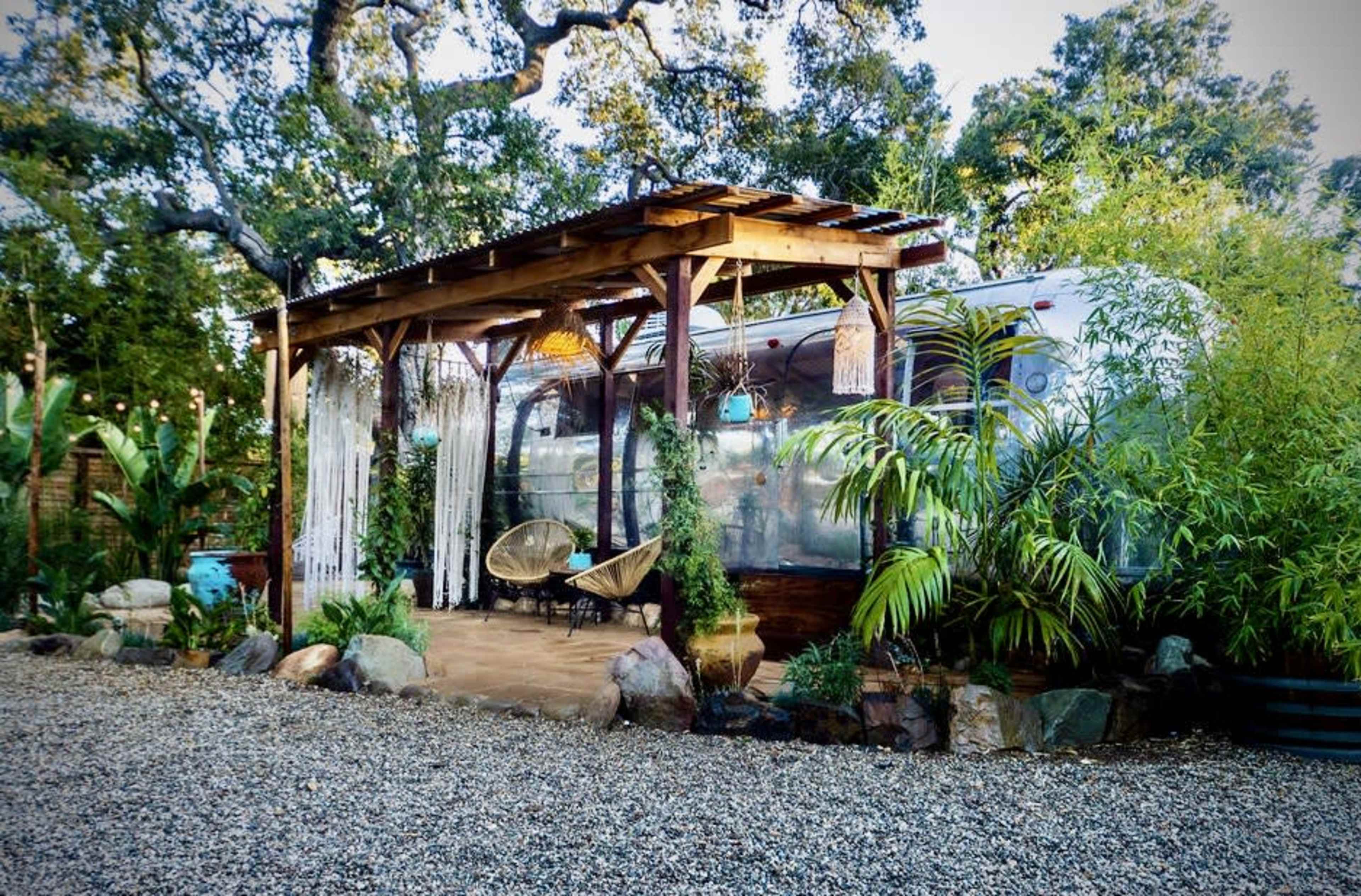 A metal trailer is positioned under a wooden pergola, surrounded by lush greenery and decorative stones in a gravel setting.