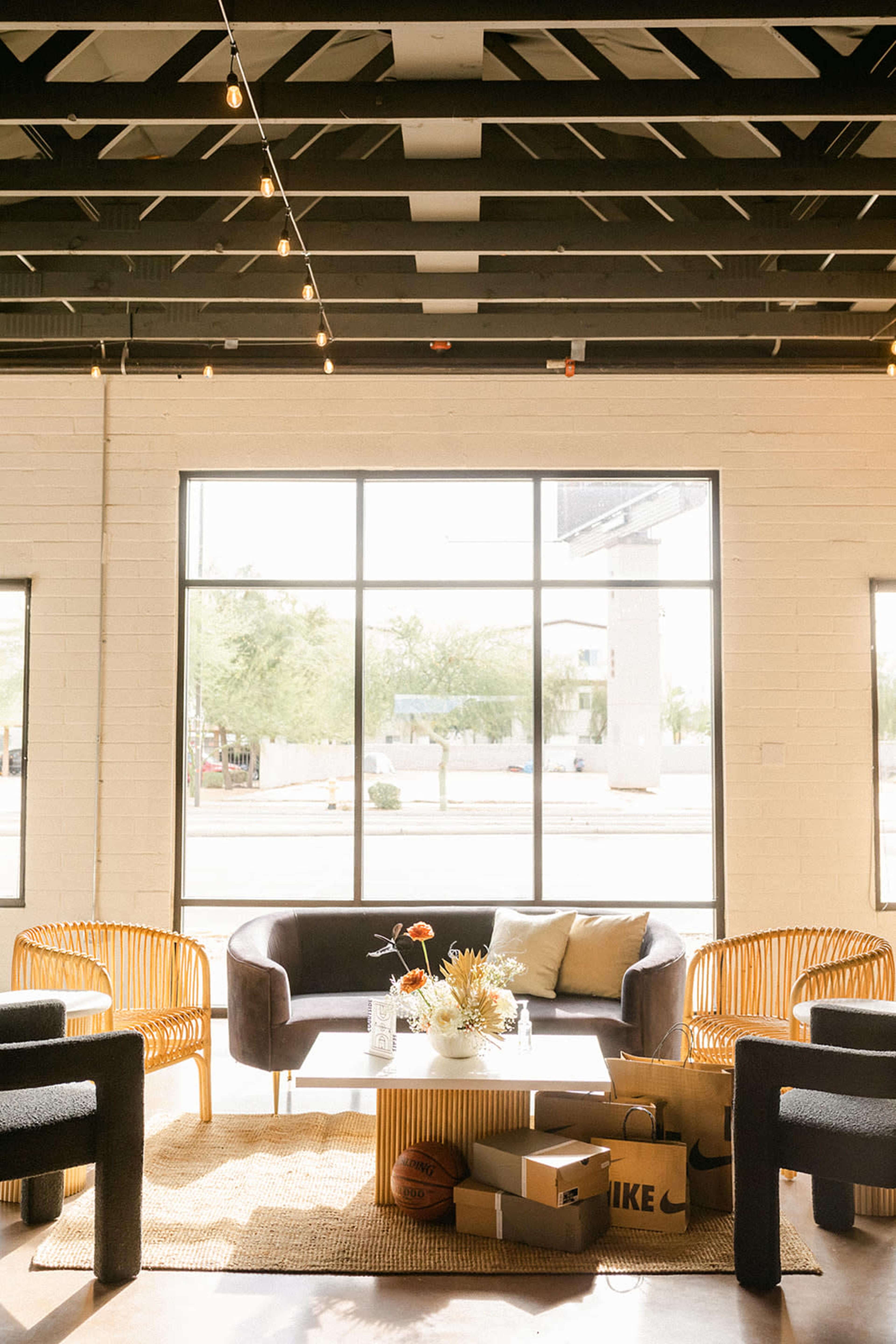 The image shows a bright indoor lounge area with modern furniture, including a dark sofa and wicker chairs, set against large windows that provide a view of the outdoors.