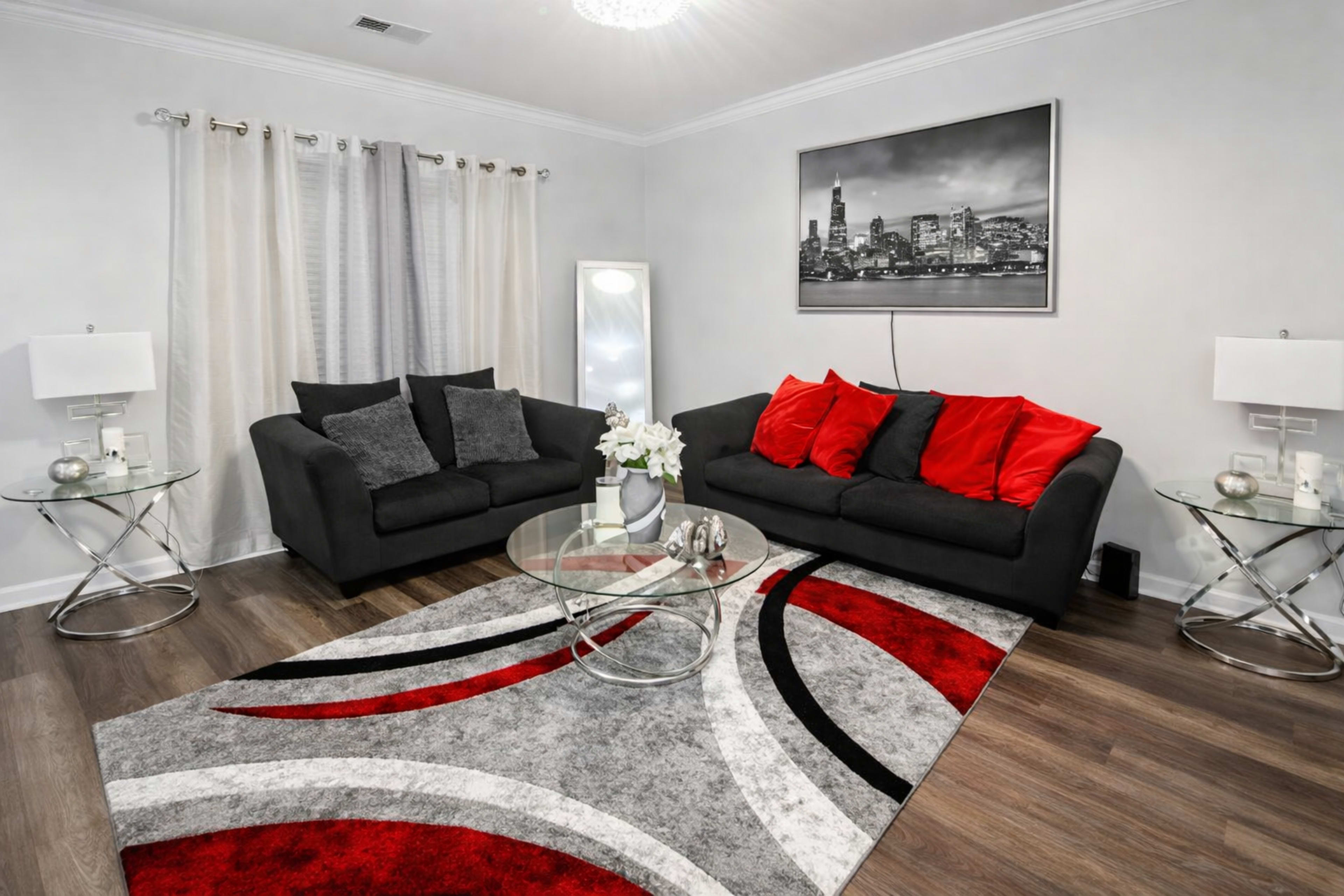 The image shows a modern living room with a black sofa set, red accent pillows, a round glass coffee table, and a gray and red area rug, accompanied by decorative lamps and wall art.