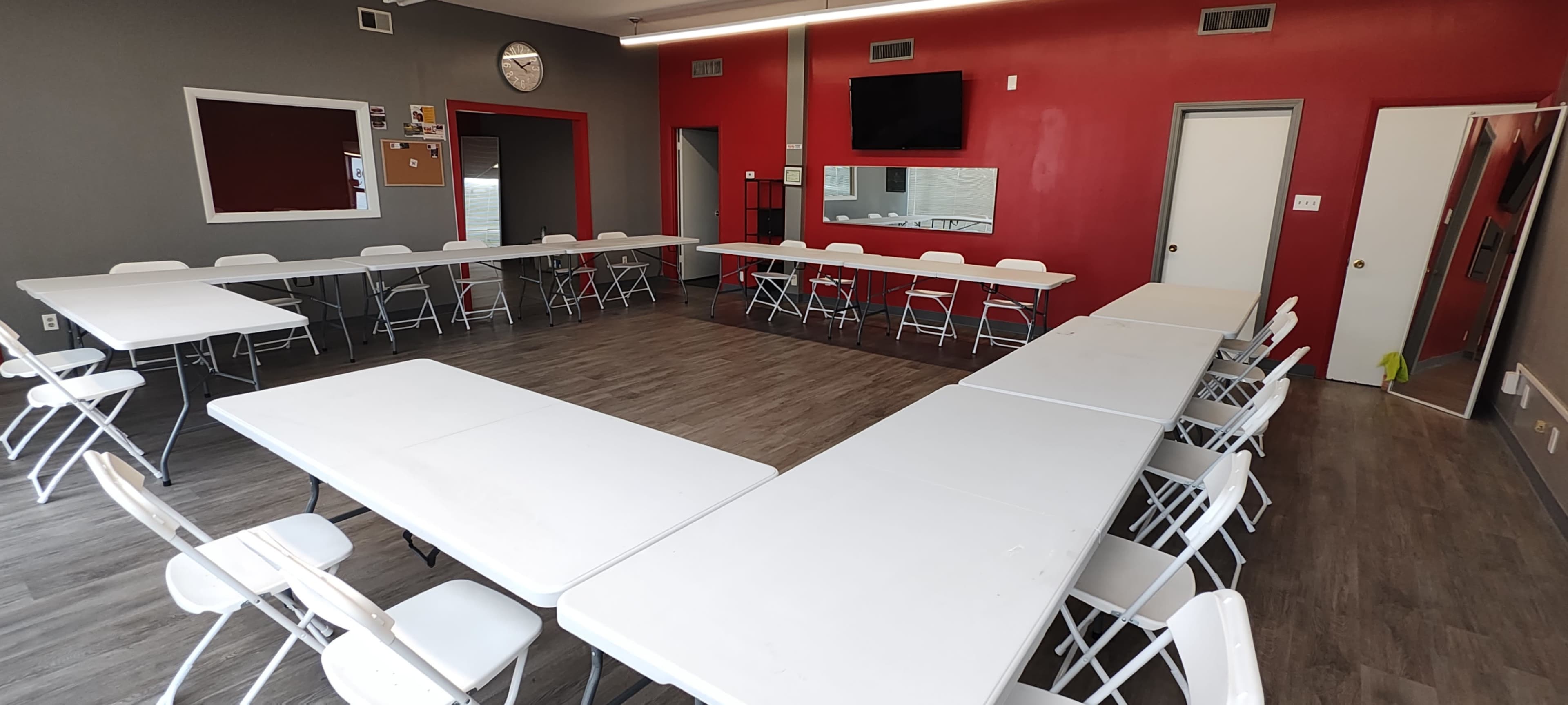 A room is set up for a meeting, featuring several white folding tables arranged in a U-shape with plastic chairs around them, alongside red and gray walls.
