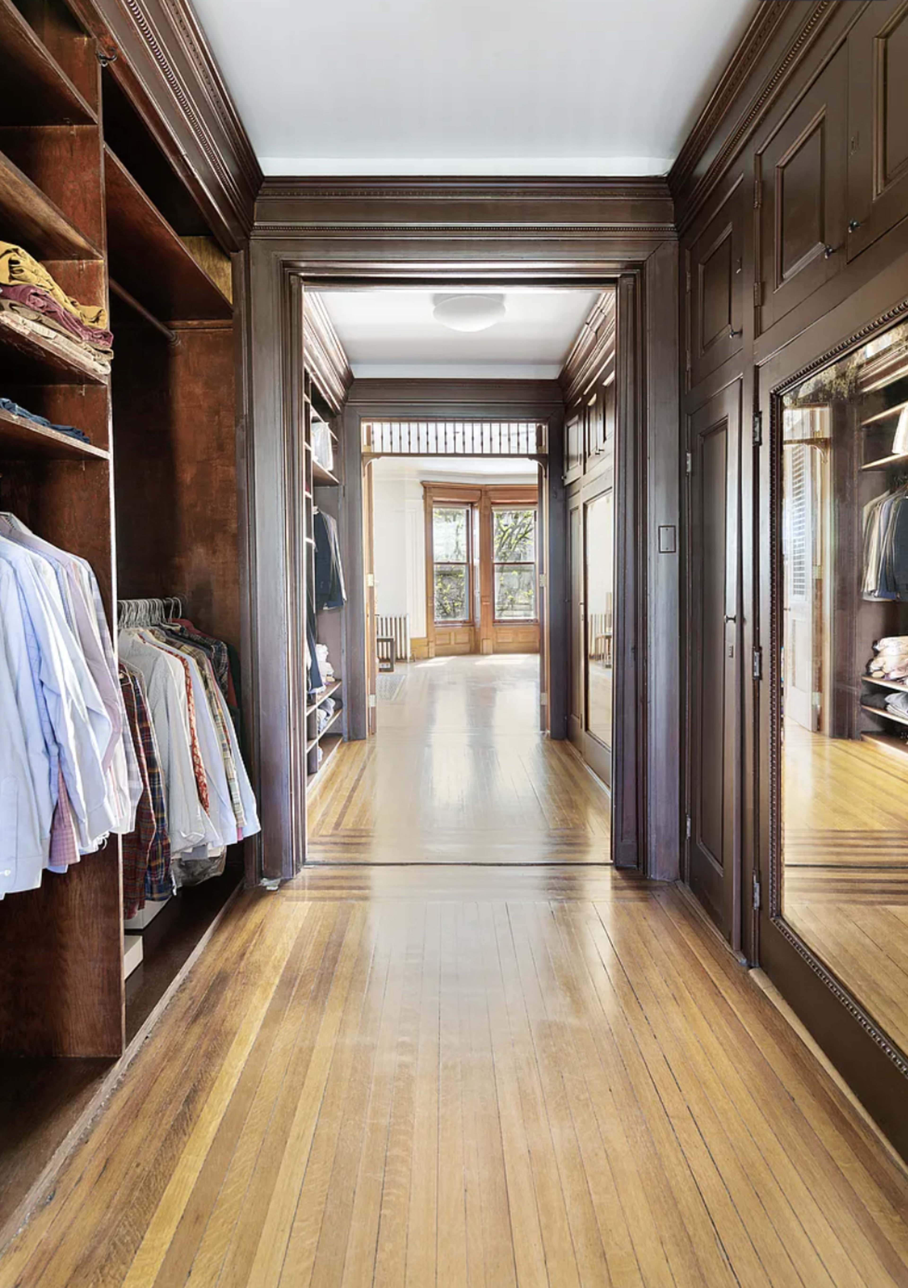 A spacious hallway features a large closet with neatly arranged clothes and a mirrored wall reflecting the light from the adjoining room.