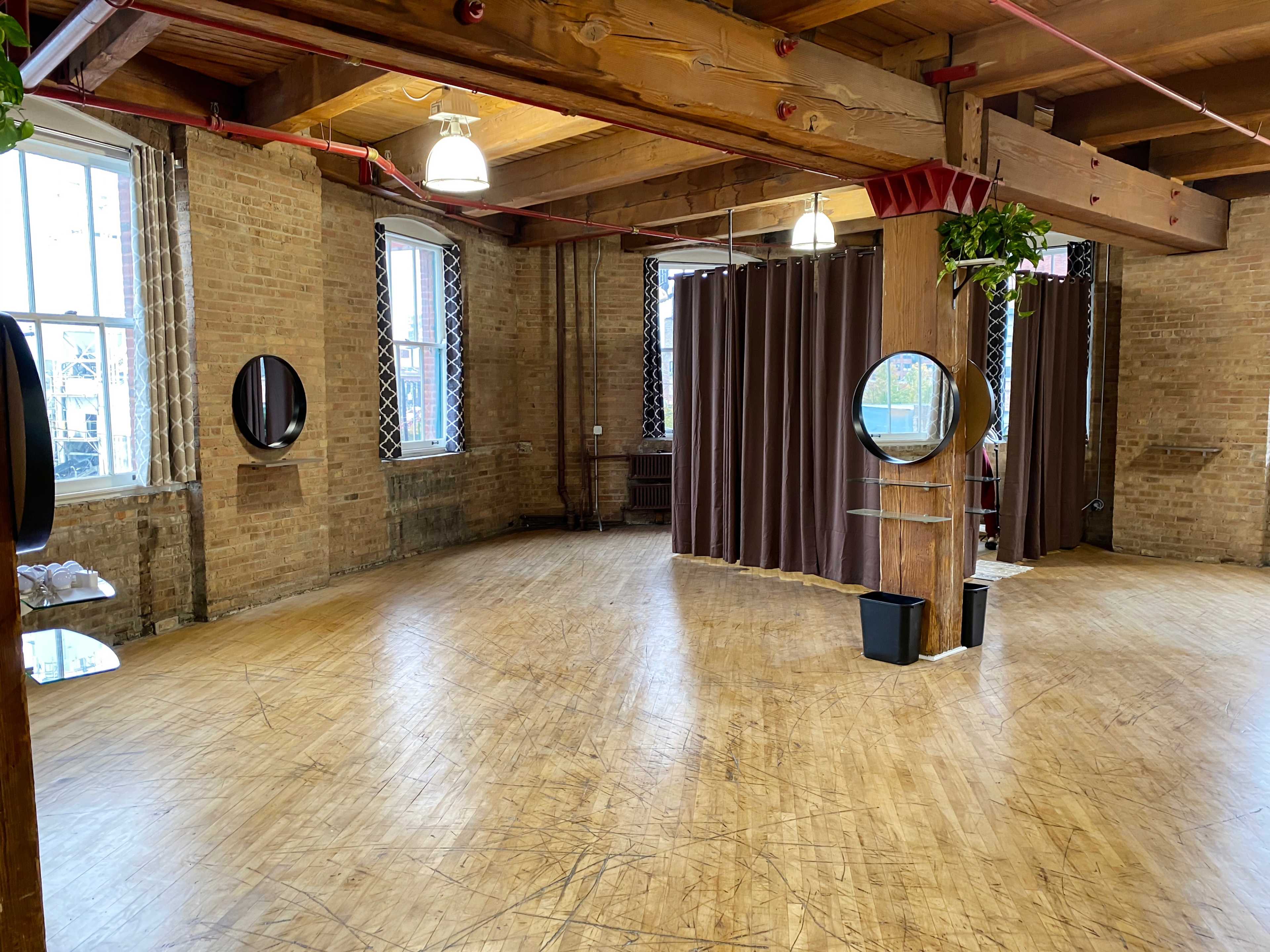 The image shows a spacious, well-lit room featuring exposed brick walls, wooden beams, and several mirrors.