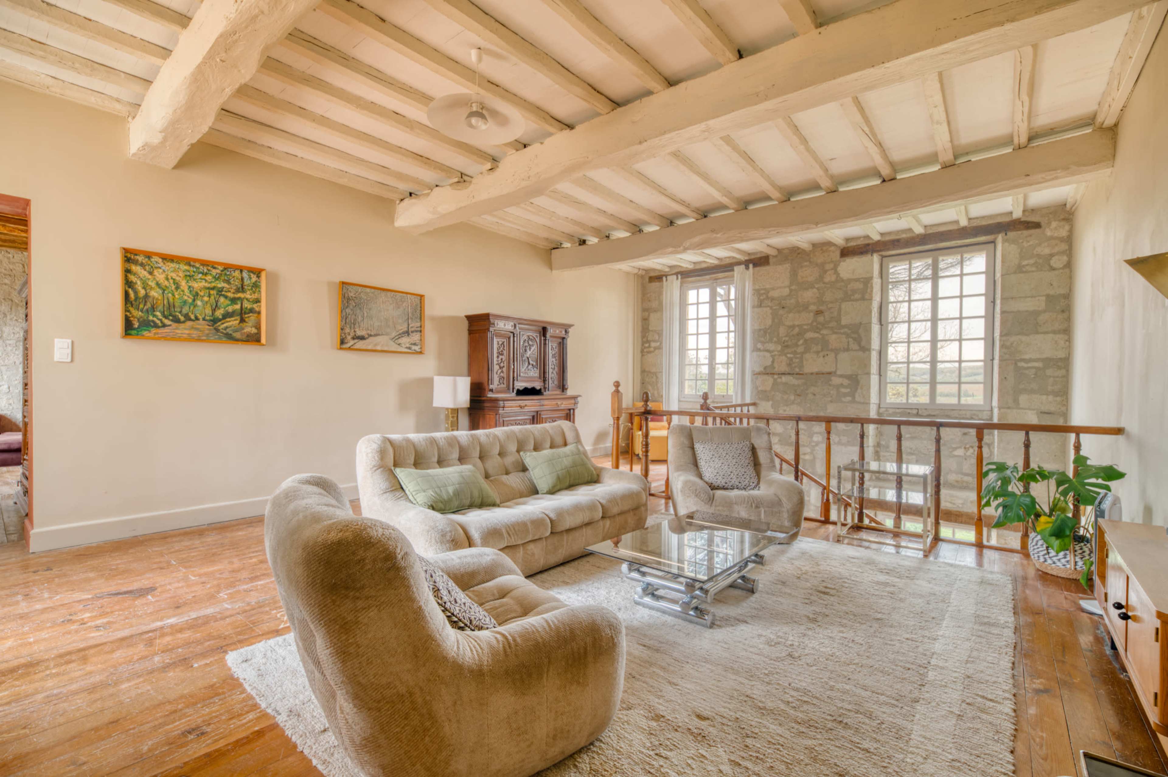 The image shows a spacious living room with wooden beams, a light-colored sofa set, a glass coffee table, and stone walls, complemented by artworks and a potted plant.
