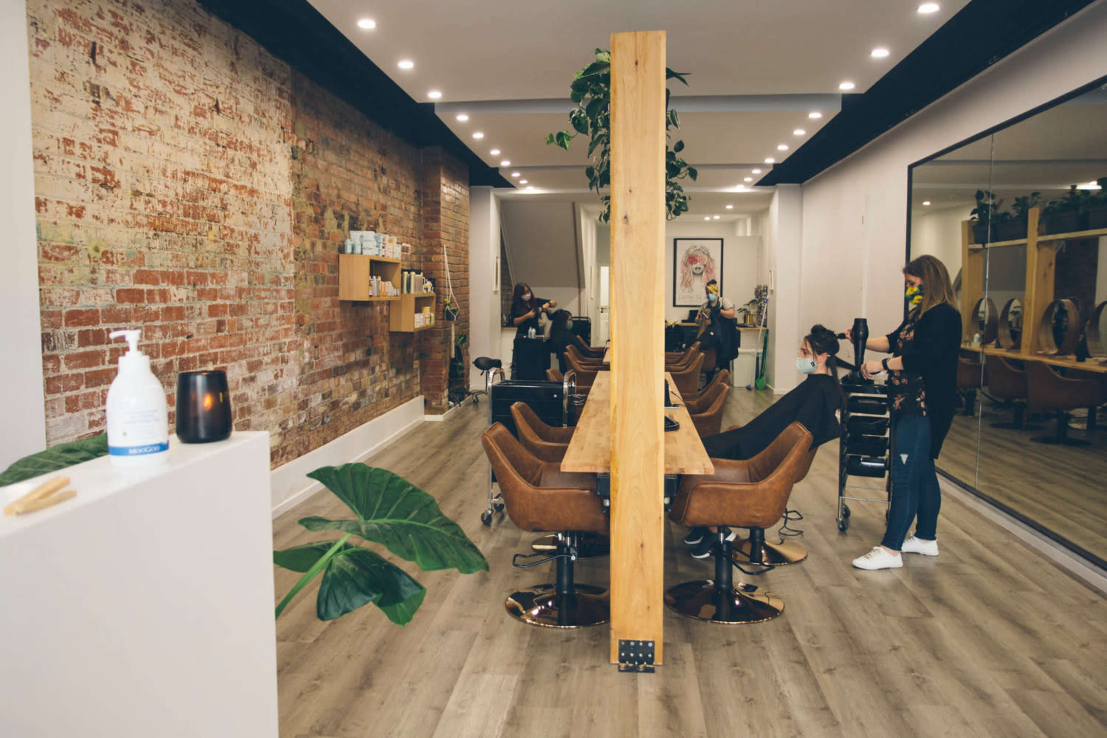 A modern hair salon features a brick wall, wooden furniture, and styling stations with clients and stylists in action.