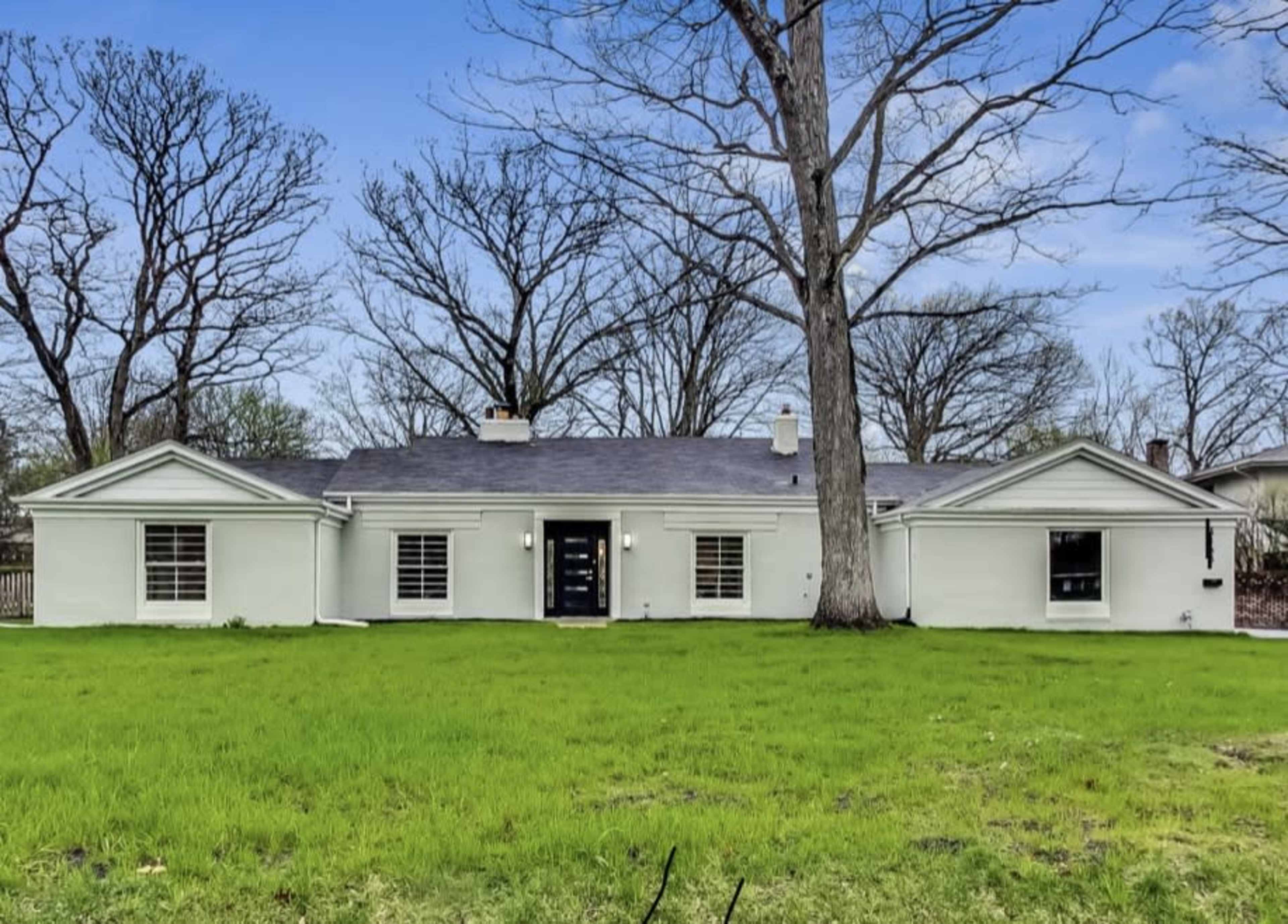 A single-story, light-colored house with a flat roof sits in a grassy yard, surrounded by bare trees.