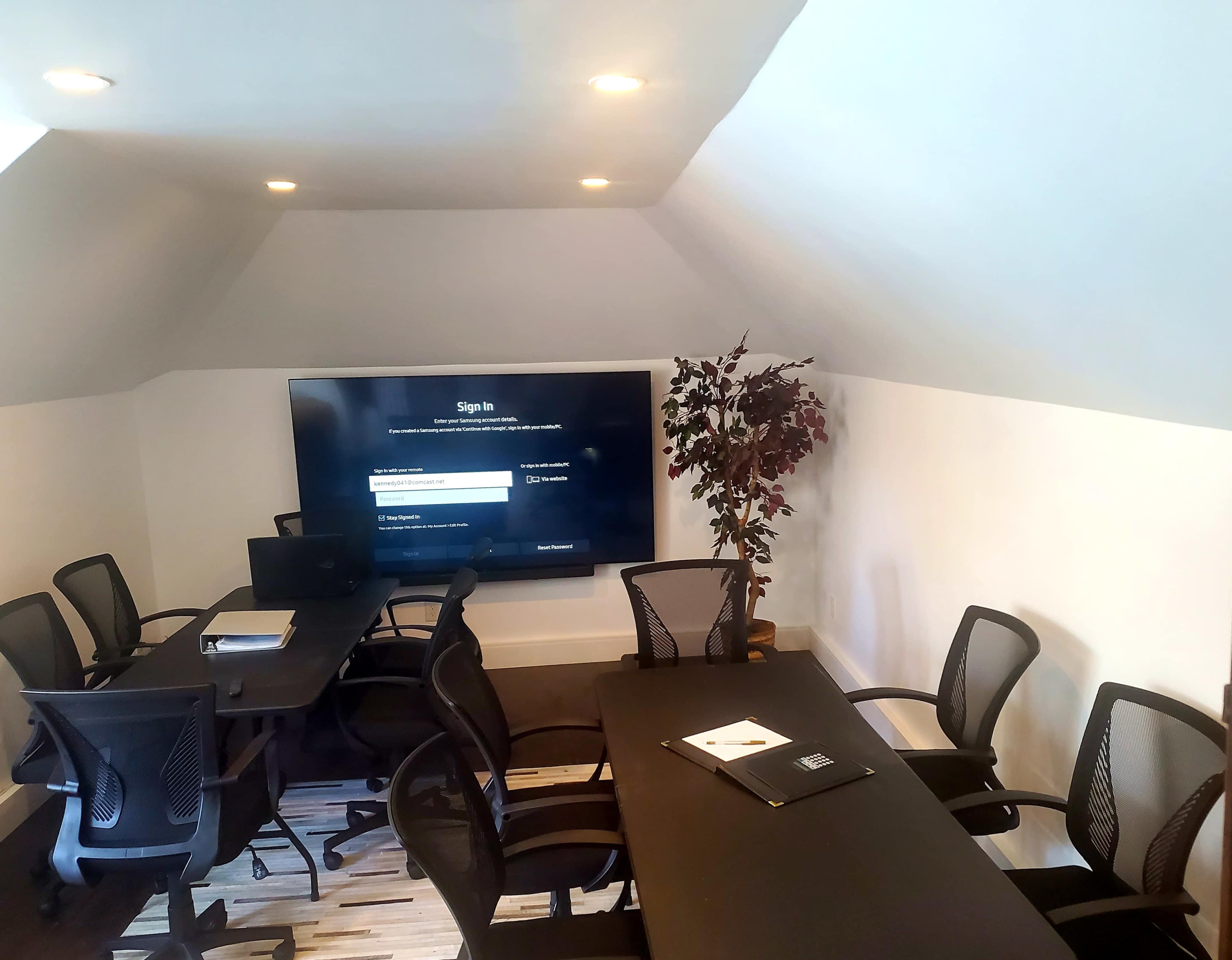 A meeting room features a large screen at one end, surrounded by several black chairs and a table, with a plant in the corner.