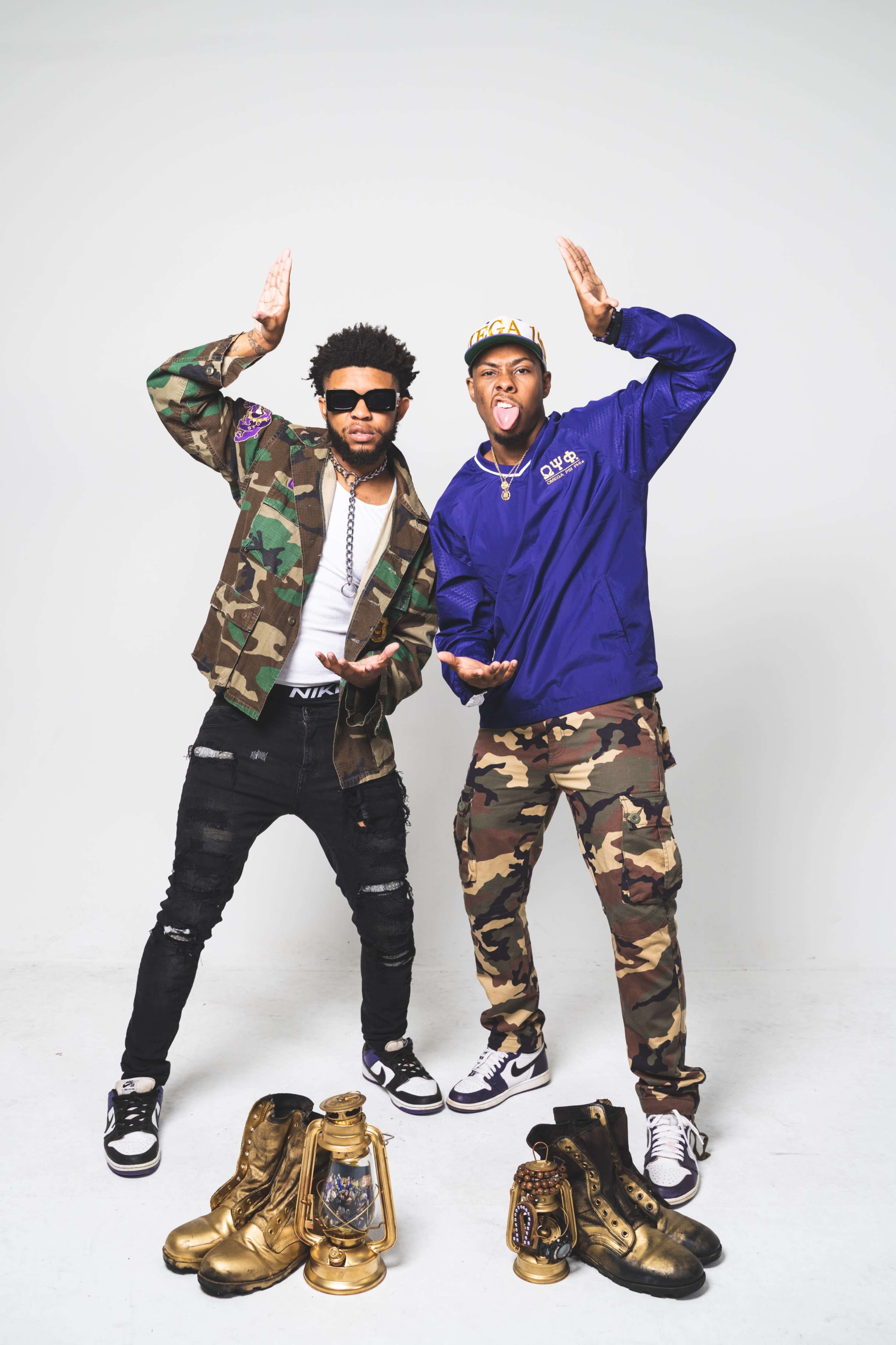 Two men pose in a studio wearing camouflage outfits, sunglasses, and sneakers, with a pair of golden boots and a vintage lantern placed on the floor in front of them.