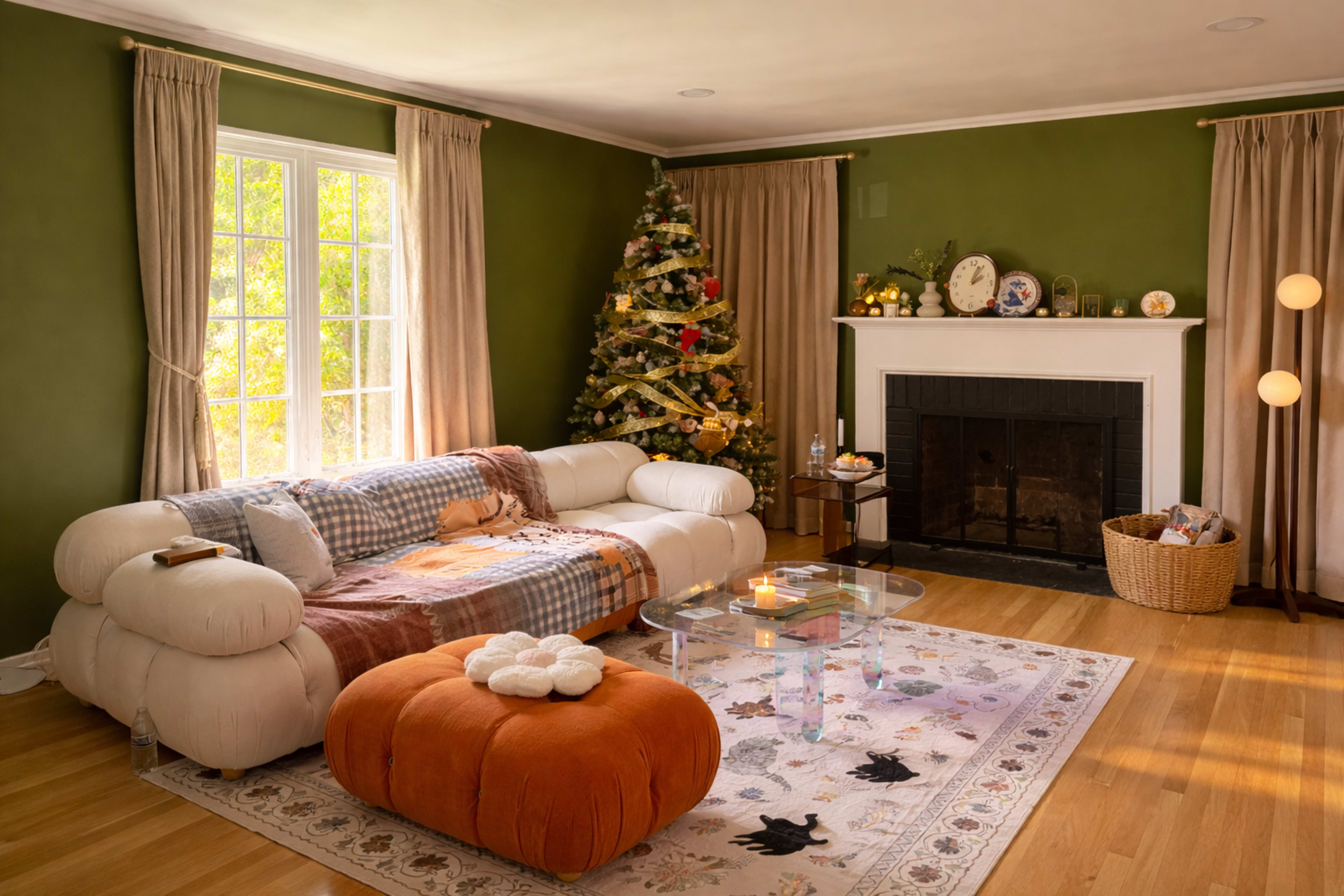 The image shows a cozy living room featuring a white sectional sofa, a decorated Christmas tree, a fireplace with shelves, and a round orange pouf.
