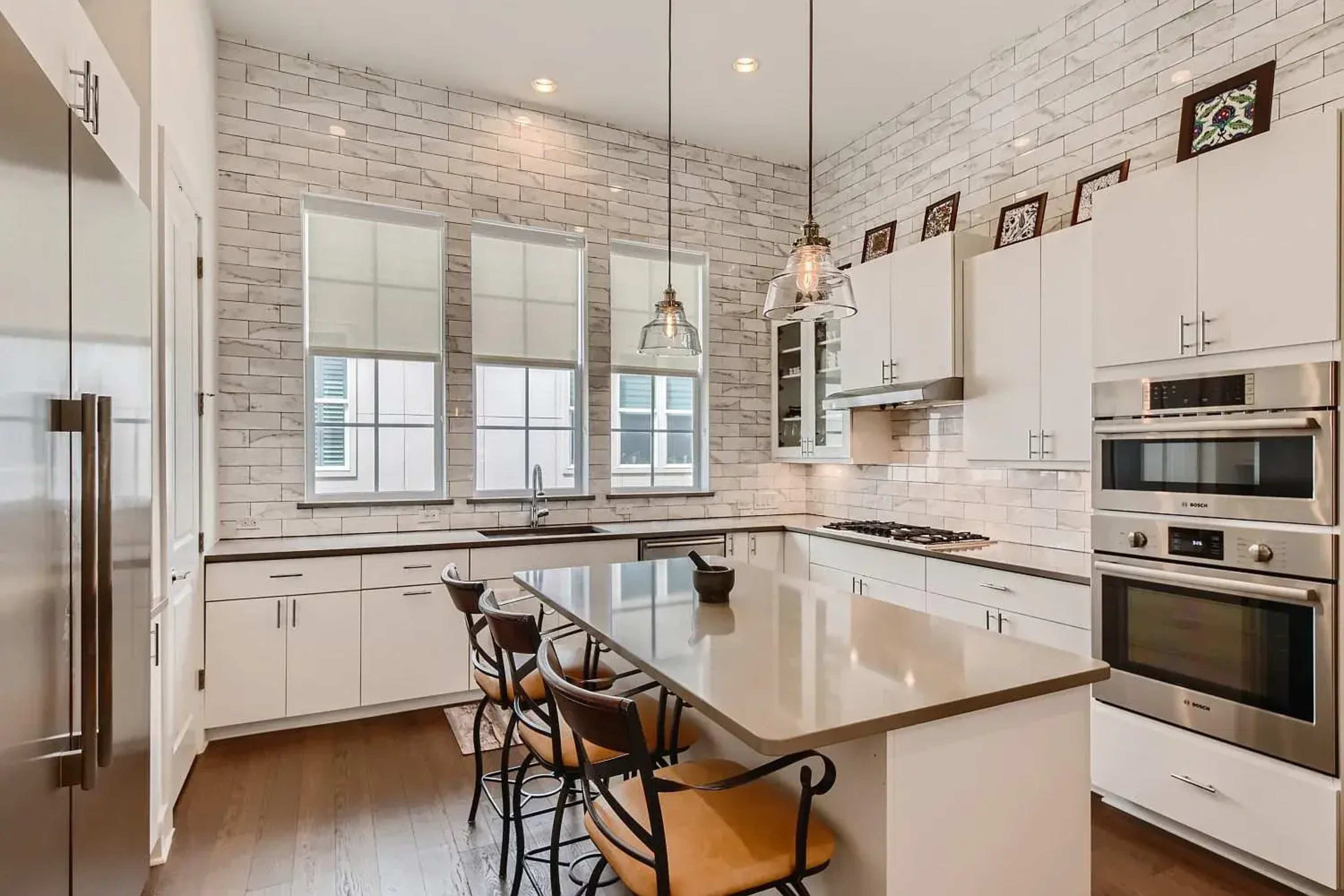 The image shows a modern kitchen with white cabinetry, a large island, stainless steel appliances, and tile backsplash.