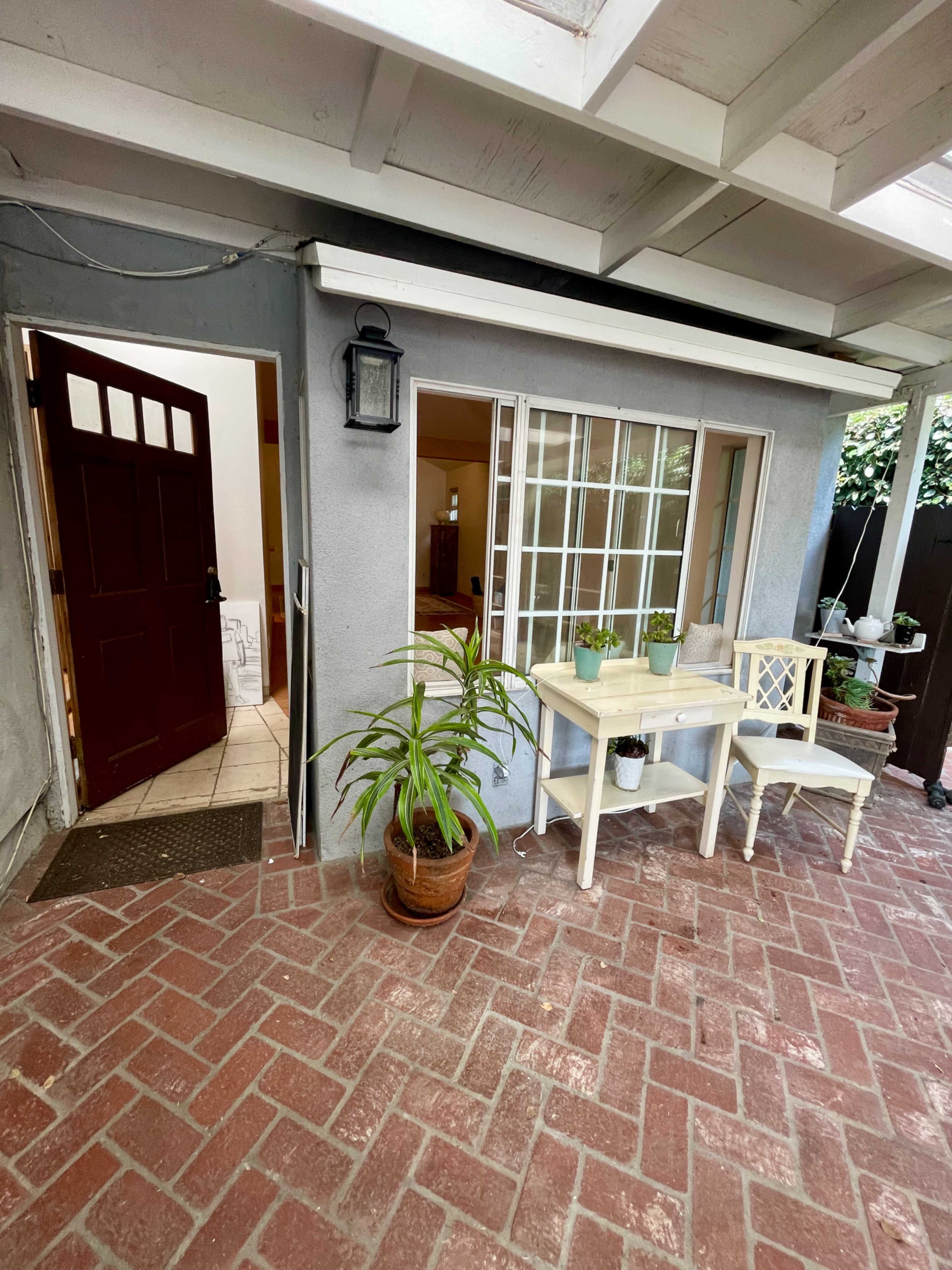 The image shows a patio area with a small table and chairs next to a door leading into a house, surrounded by potted plants and a brick floor.