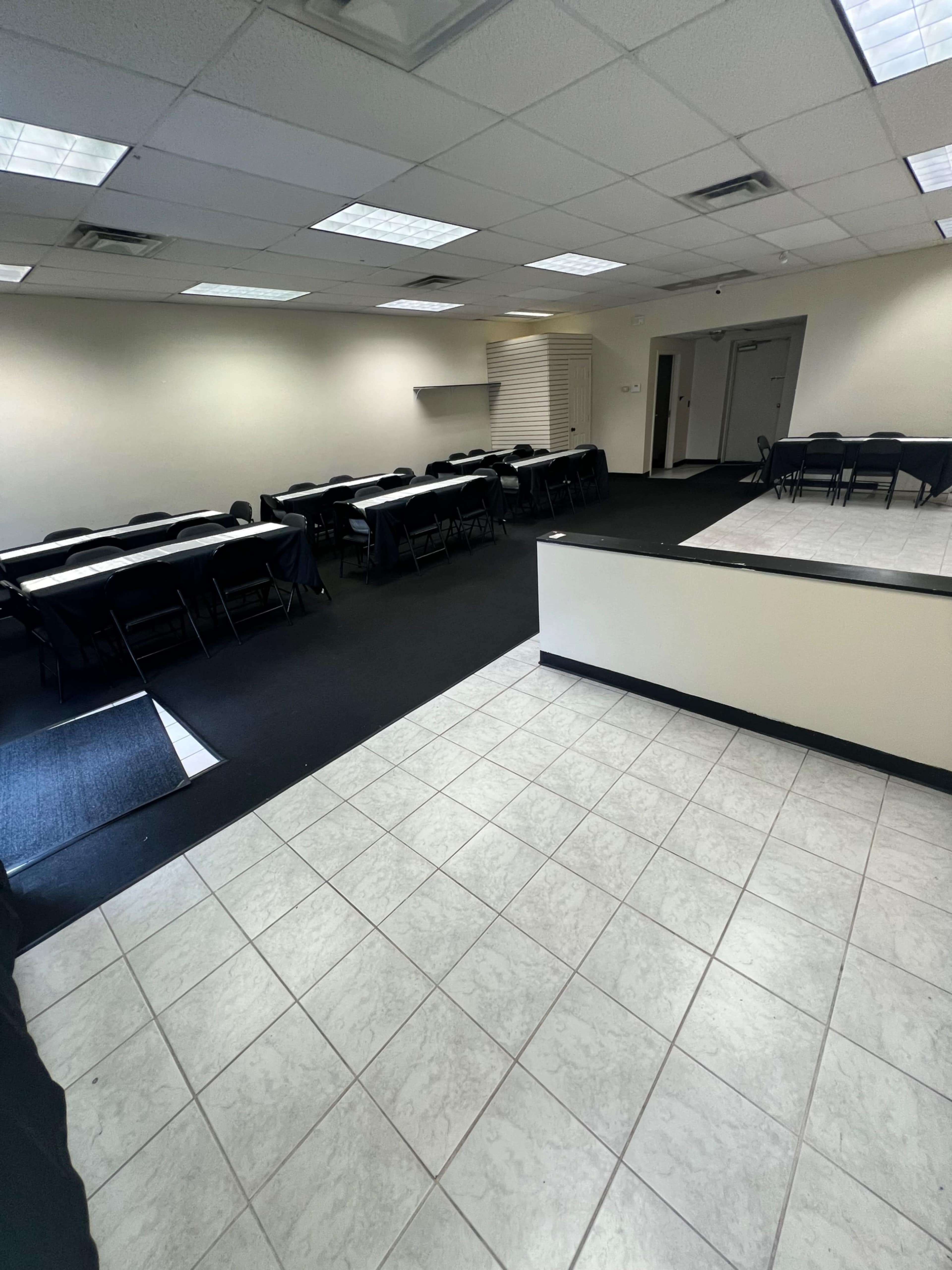 The image shows an empty meeting room with rows of rectangular tables and chairs, and a tiled floor transitioning to carpet.