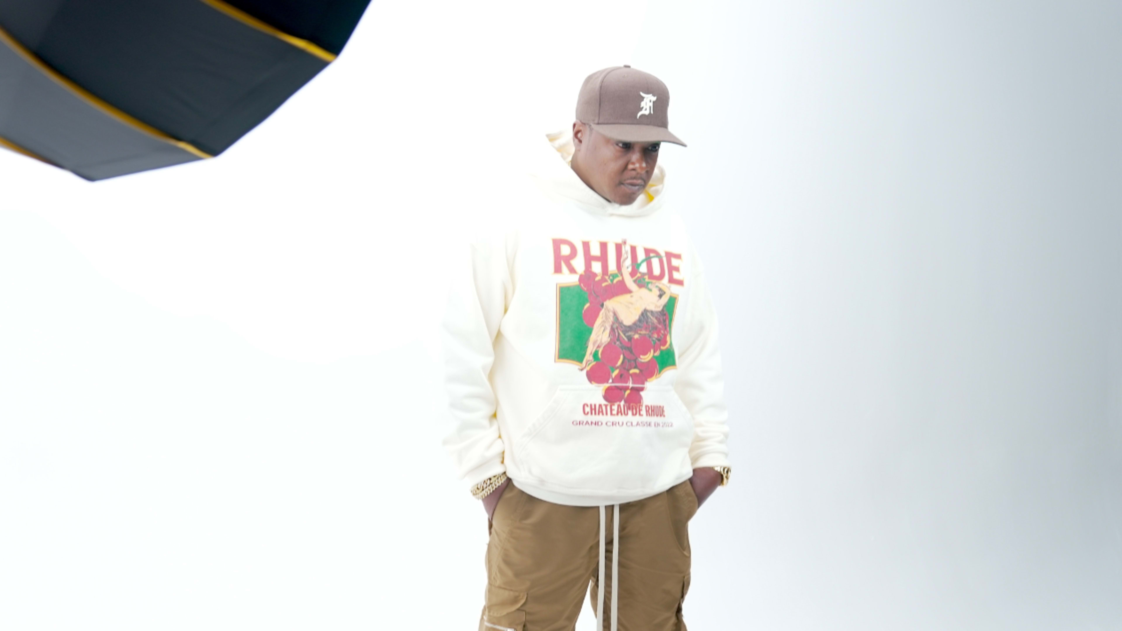 A man in a light-colored hoodie with graphics and a cap stands under a softbox light against a plain background.
