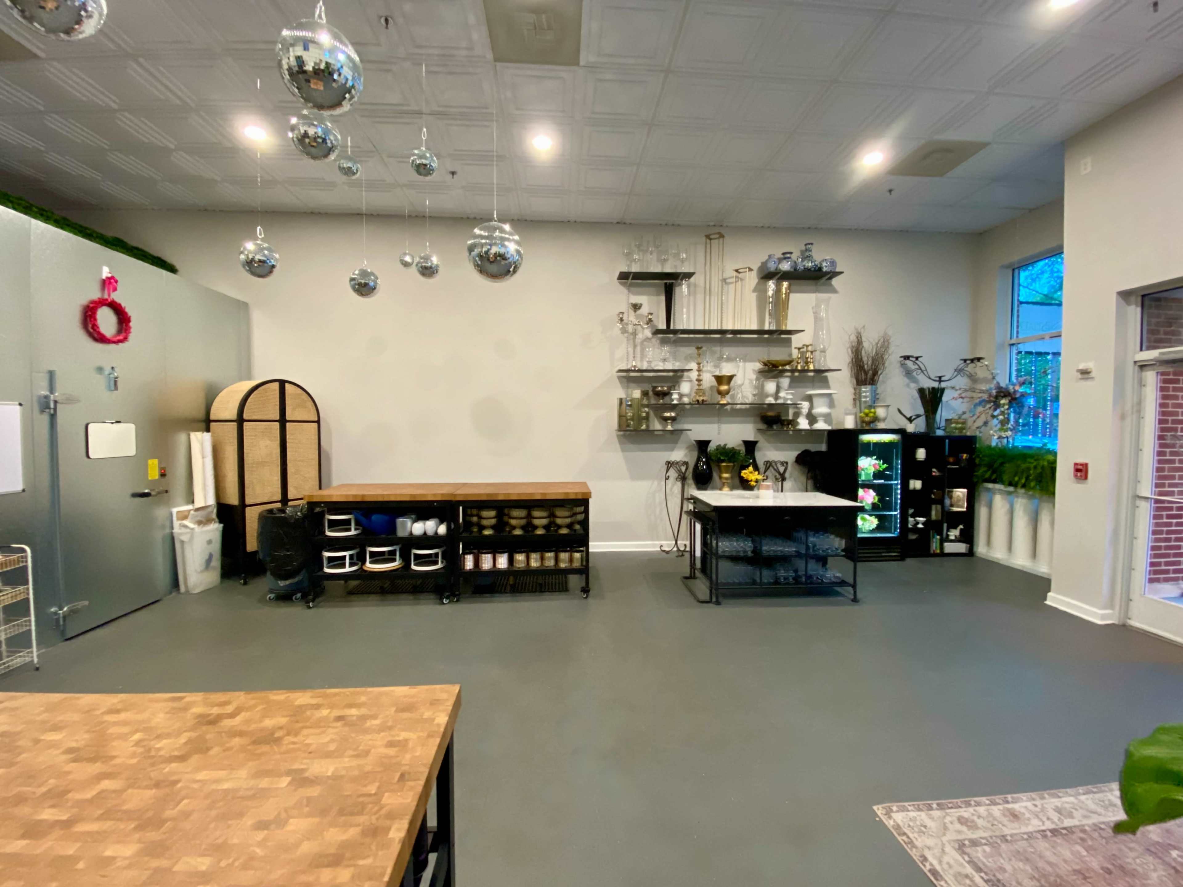 A spacious room with a large metal refrigerator, a wooden work table, open shelving displaying decorative items, and a window allowing natural light to enter.