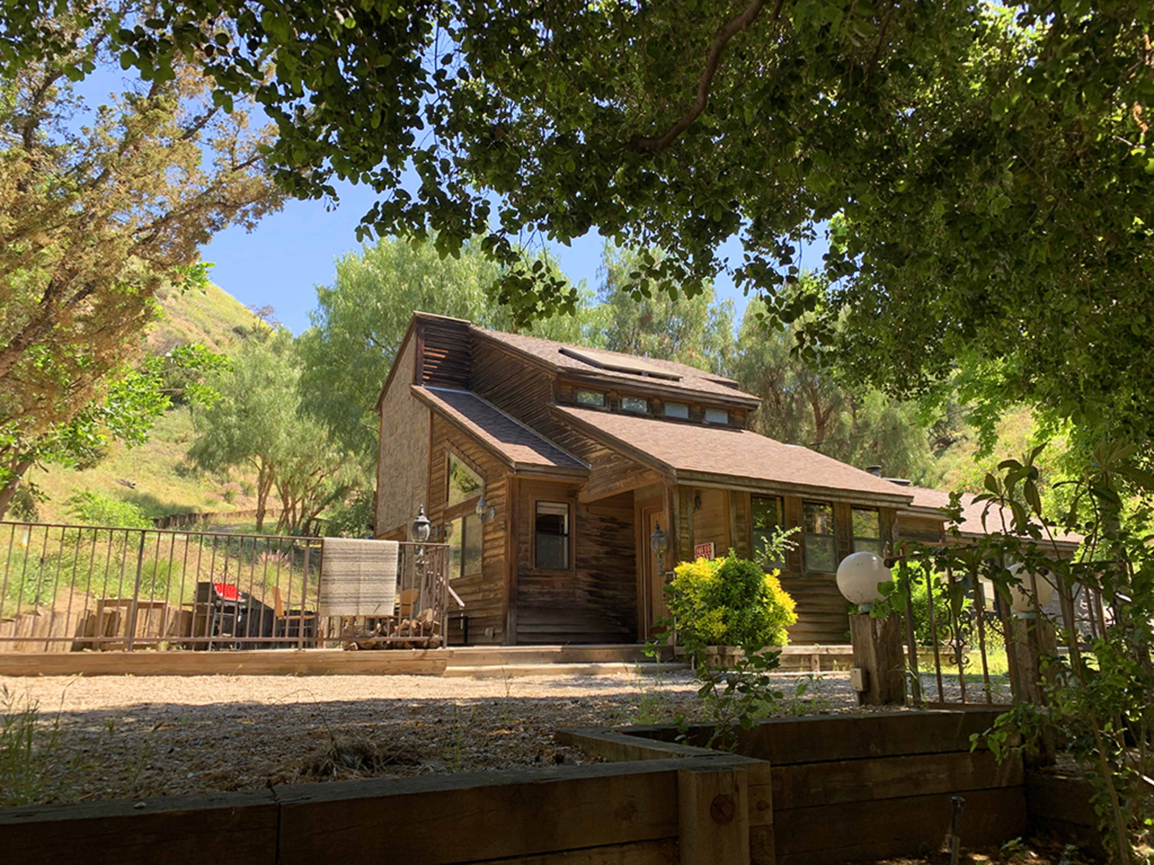 A wooden house with a sloped roof sits amidst trees on a hillside, featuring a fenced garden area in the foreground.
