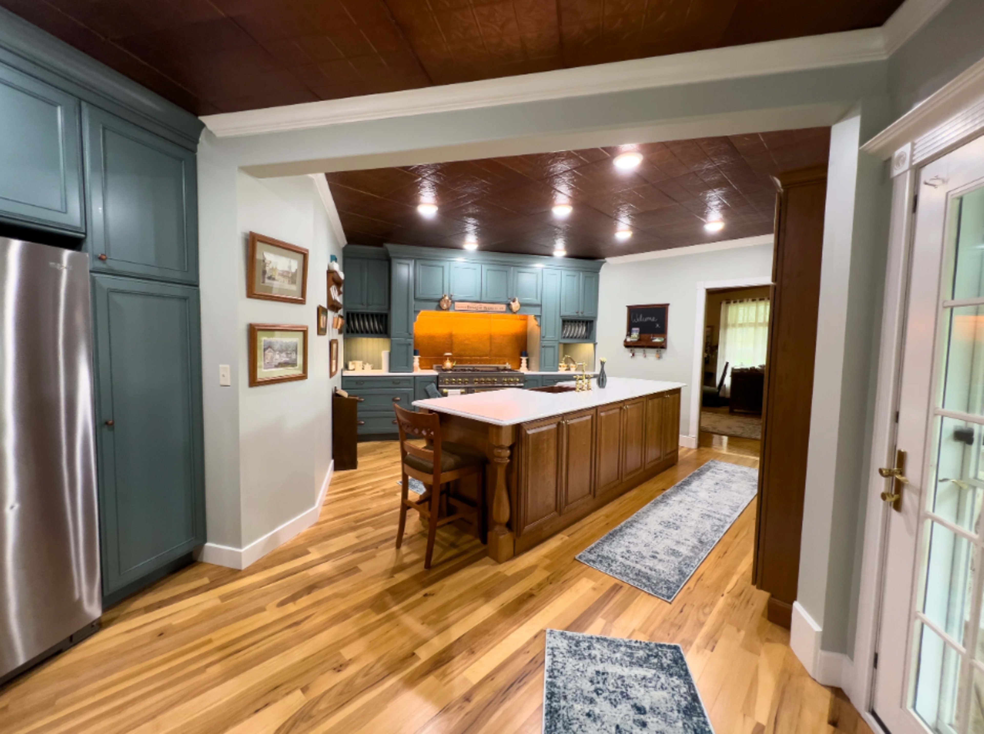 A spacious kitchen with teal cabinets, a central island with seating, hardwood flooring, and framed artwork on the walls.