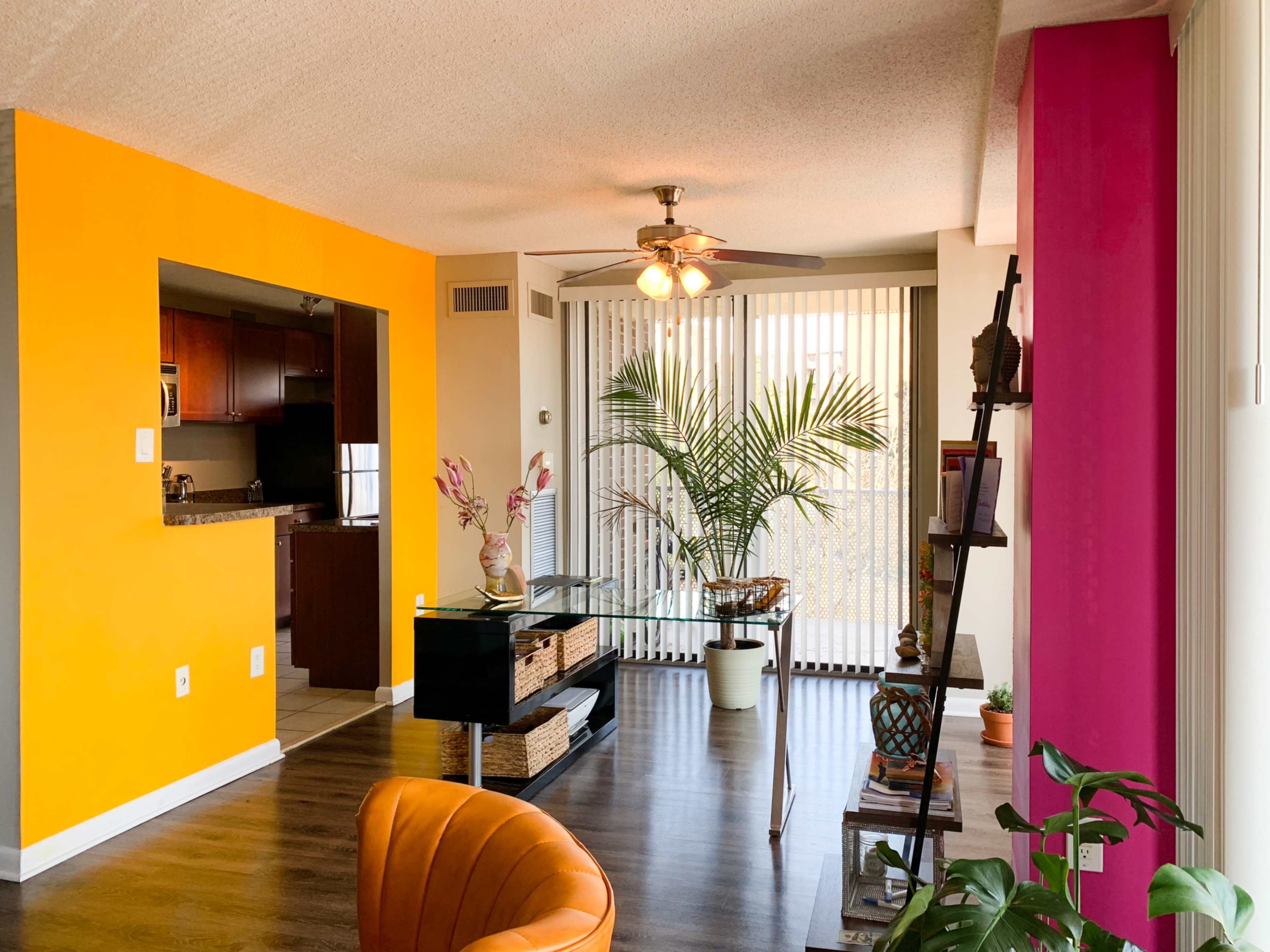A living room features bright orange and pink accent walls, a ceiling fan, a potted plant, and a modern furniture setup.
