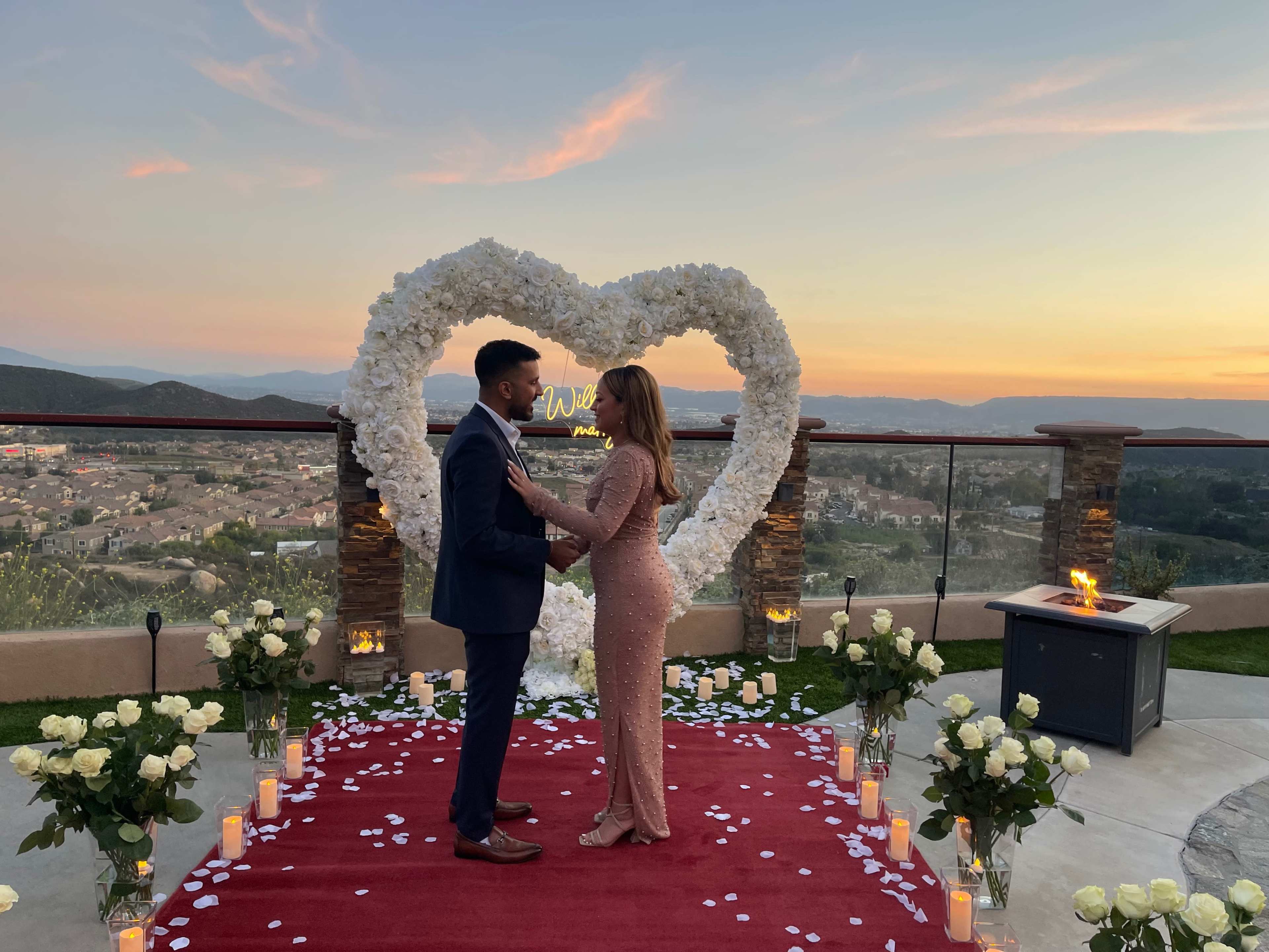 A couple stands facing each other on a red carpet adorned with rose petals, in front of a heart-shaped floral arrangement, with a scenic sunset in the background.