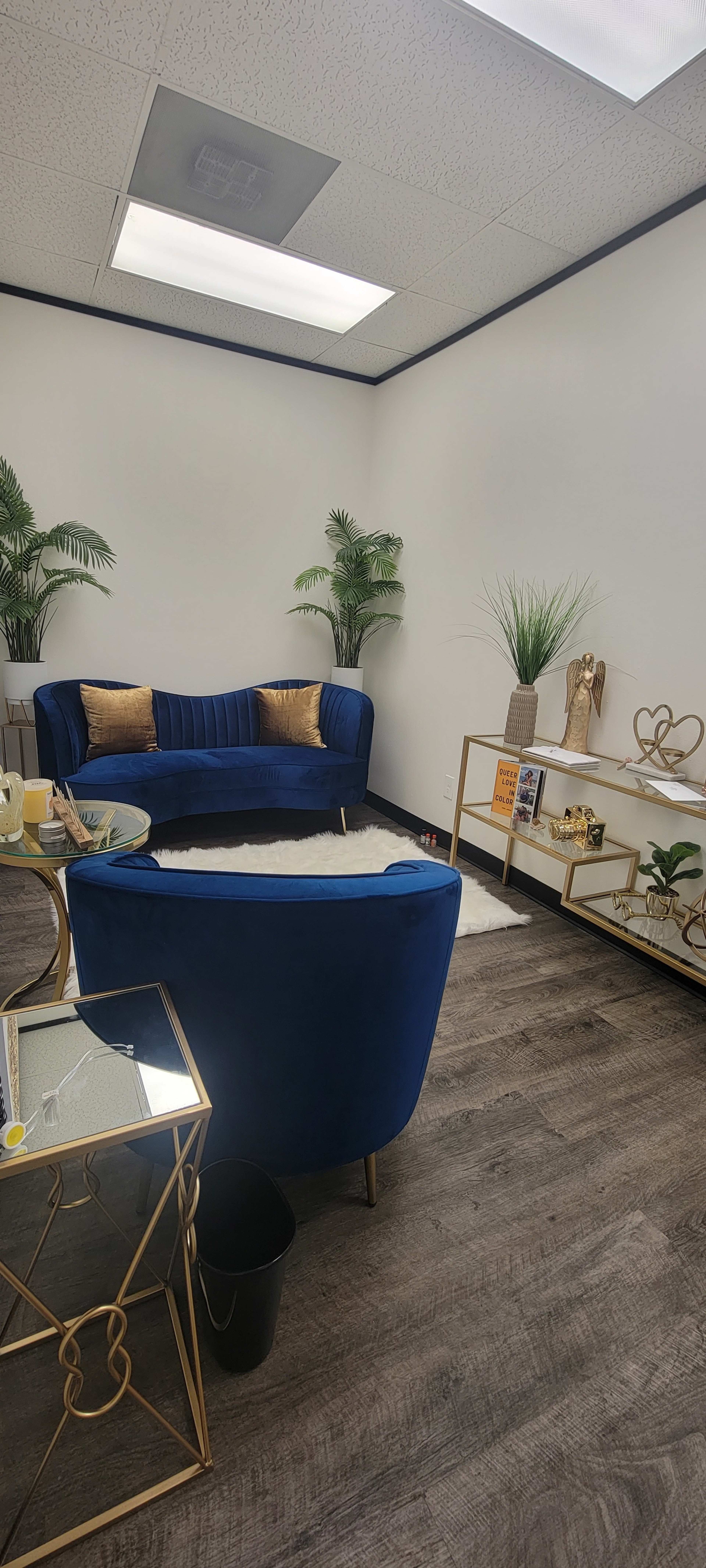A modern waiting area with a blue velvet sofa, a circular coffee table, and a decorative shelf, surrounded by potted plants.