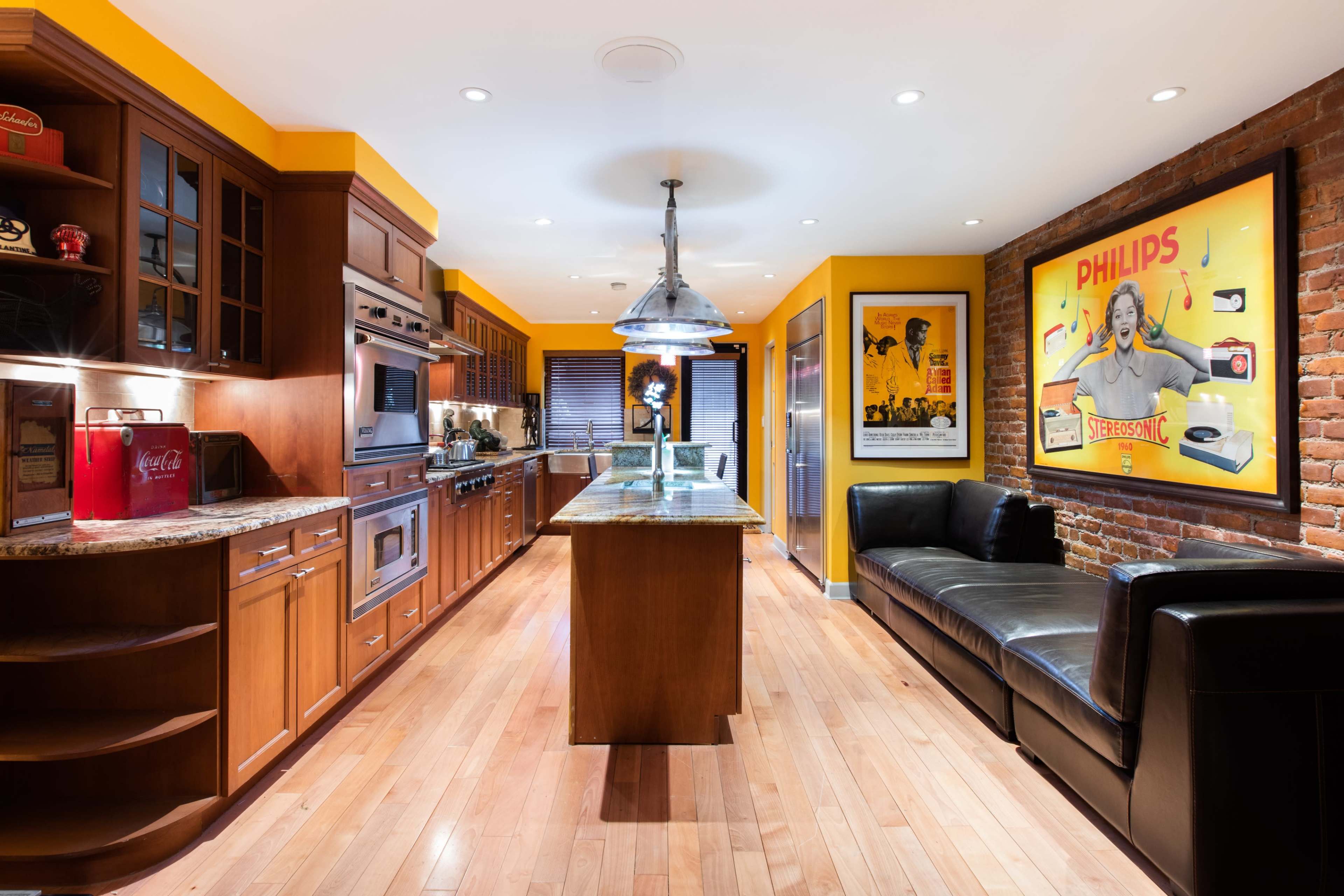 A kitchen with wooden cabinetry, a granite island, and a yellow and red color scheme, featuring a brick wall and vintage posters.