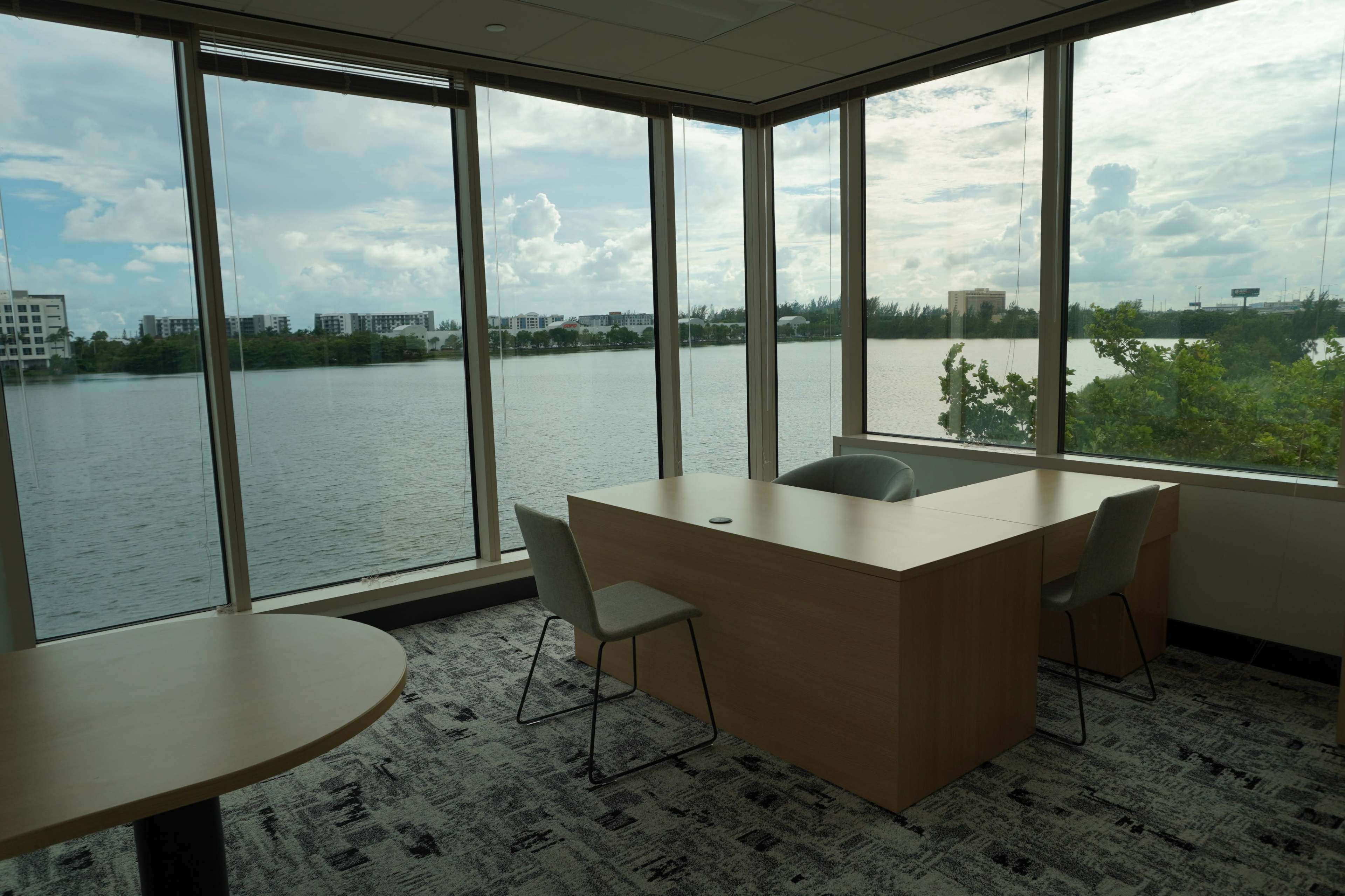 The image shows a modern office space with large windows overlooking a body of water, featuring a wooden table and chairs.