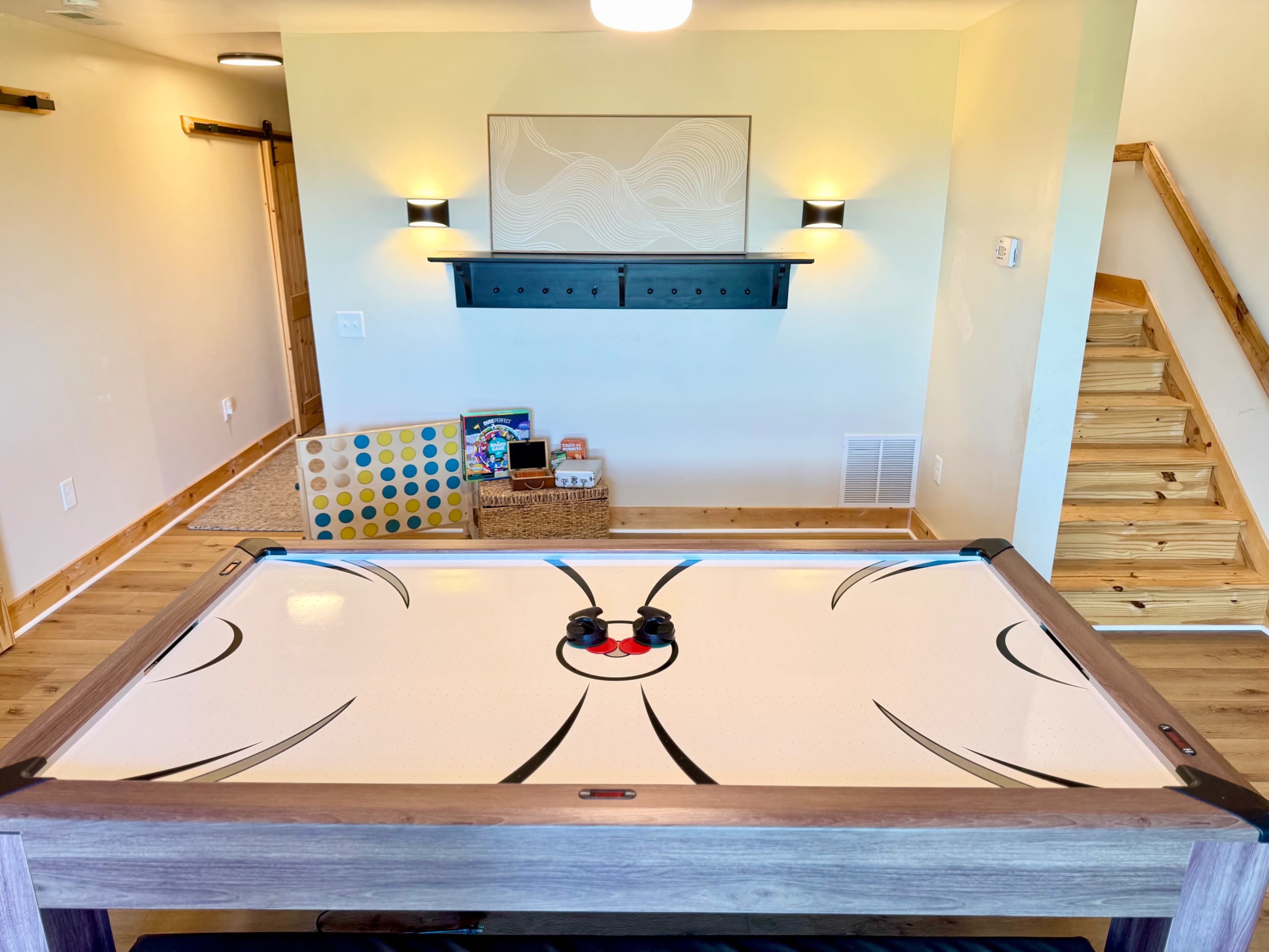 A game table is positioned in a well-lit room with wooden flooring, shelves on the wall, and a staircase in the background.