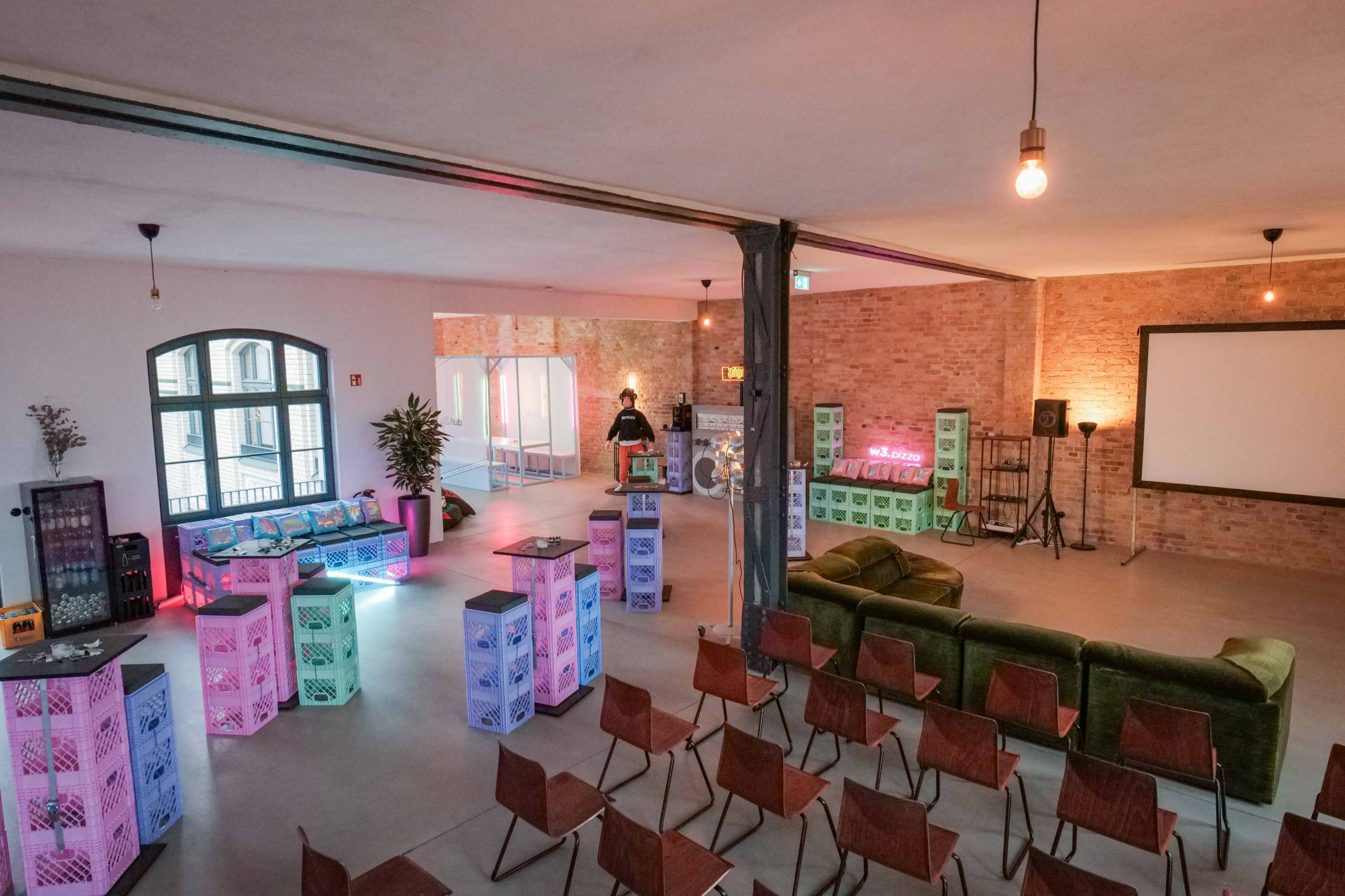 A spacious indoor venue features a mix of colorful seating areas, crates, and a projector screen, designed for social gatherings or events.