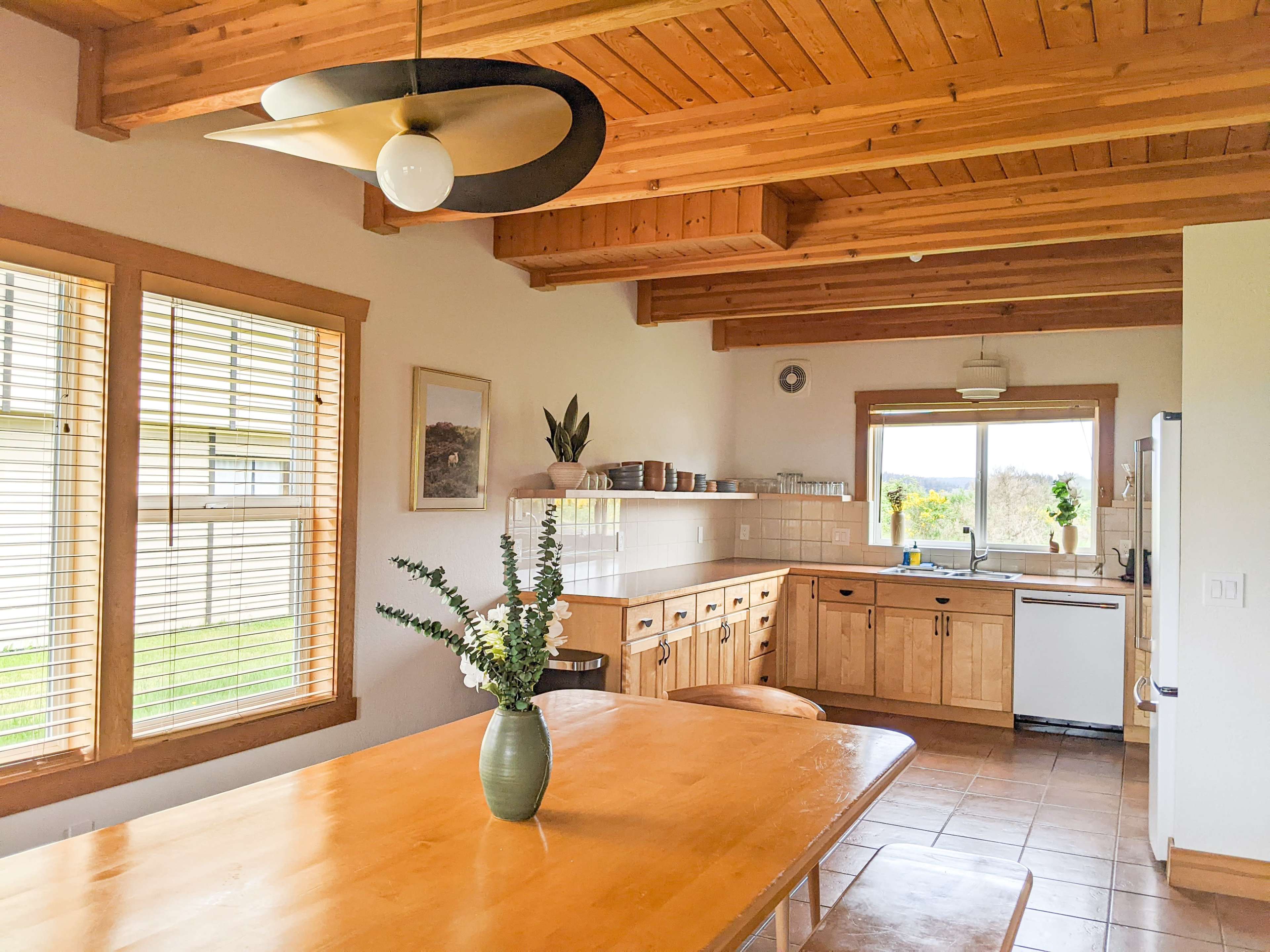 A well-lit kitchen featuring wooden cabinetry, a dining table, and large windows with blinds.