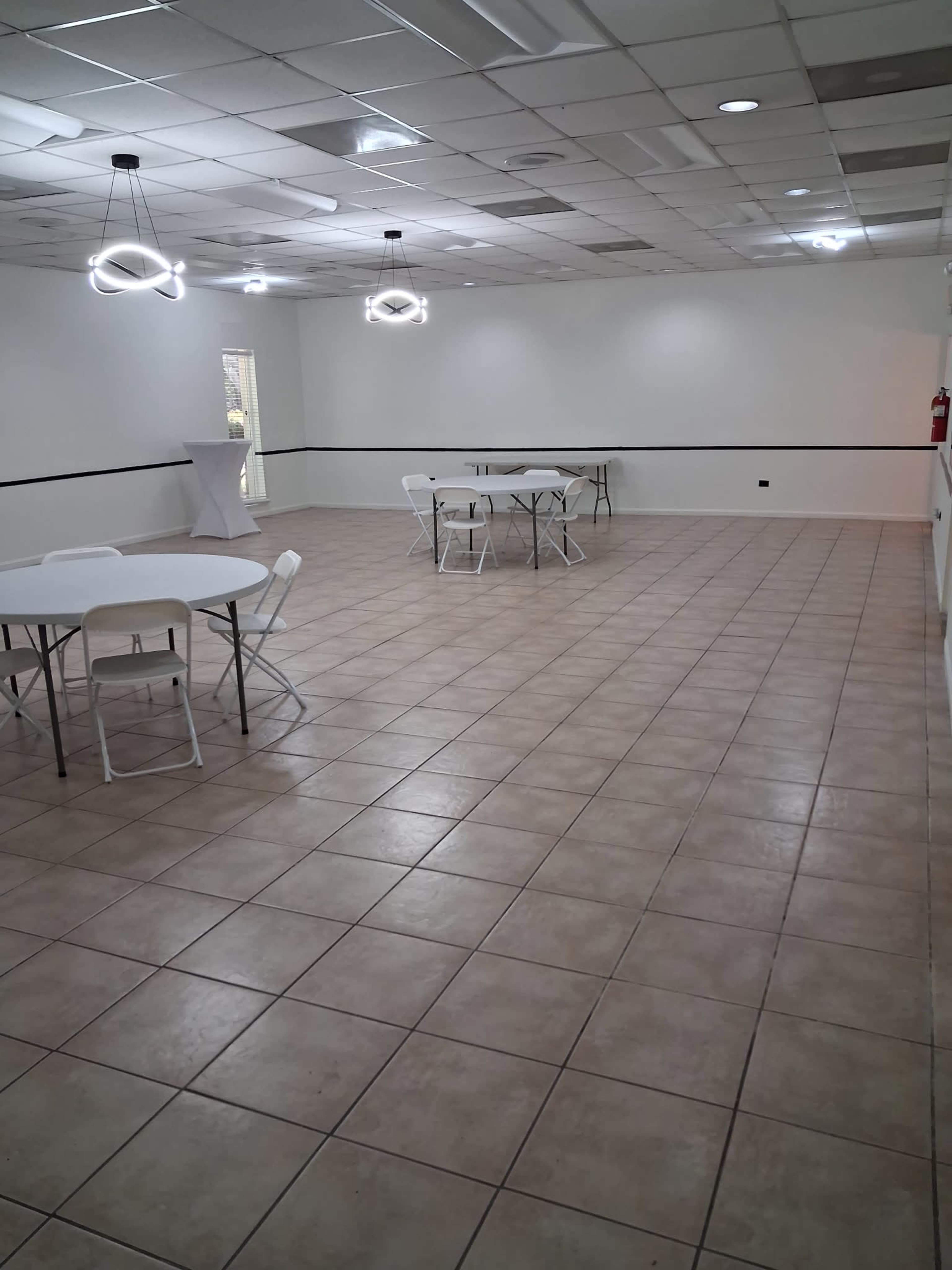 The image shows a spacious indoor area with white walls, tiled flooring, and several round tables set up with chairs.