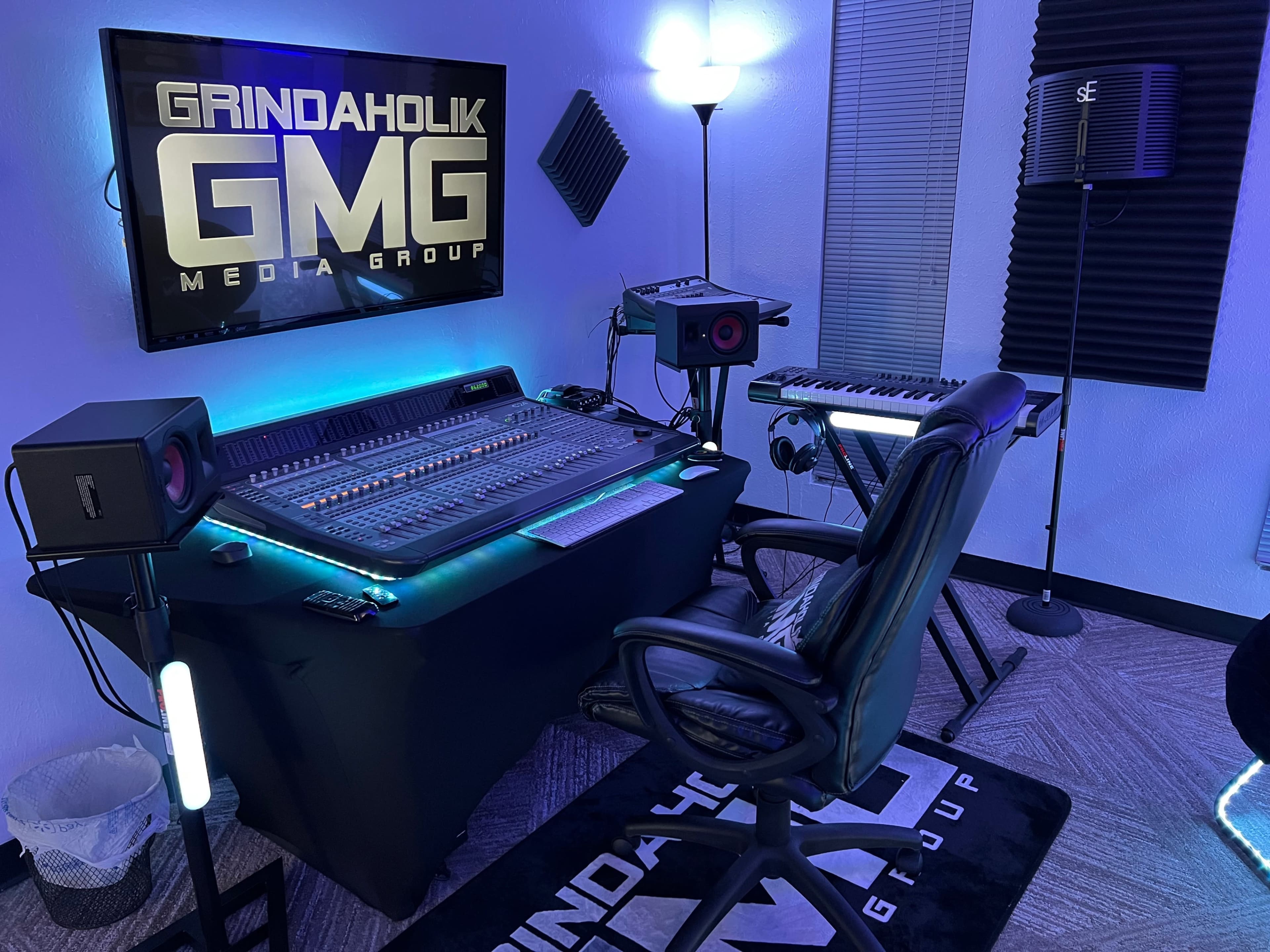 A music production studio featuring a large mixing console, a computer monitor displaying "GRINDAHOLIK GMG MEDIA GROUP," and various audio equipment, all set in a room with blue lighting.