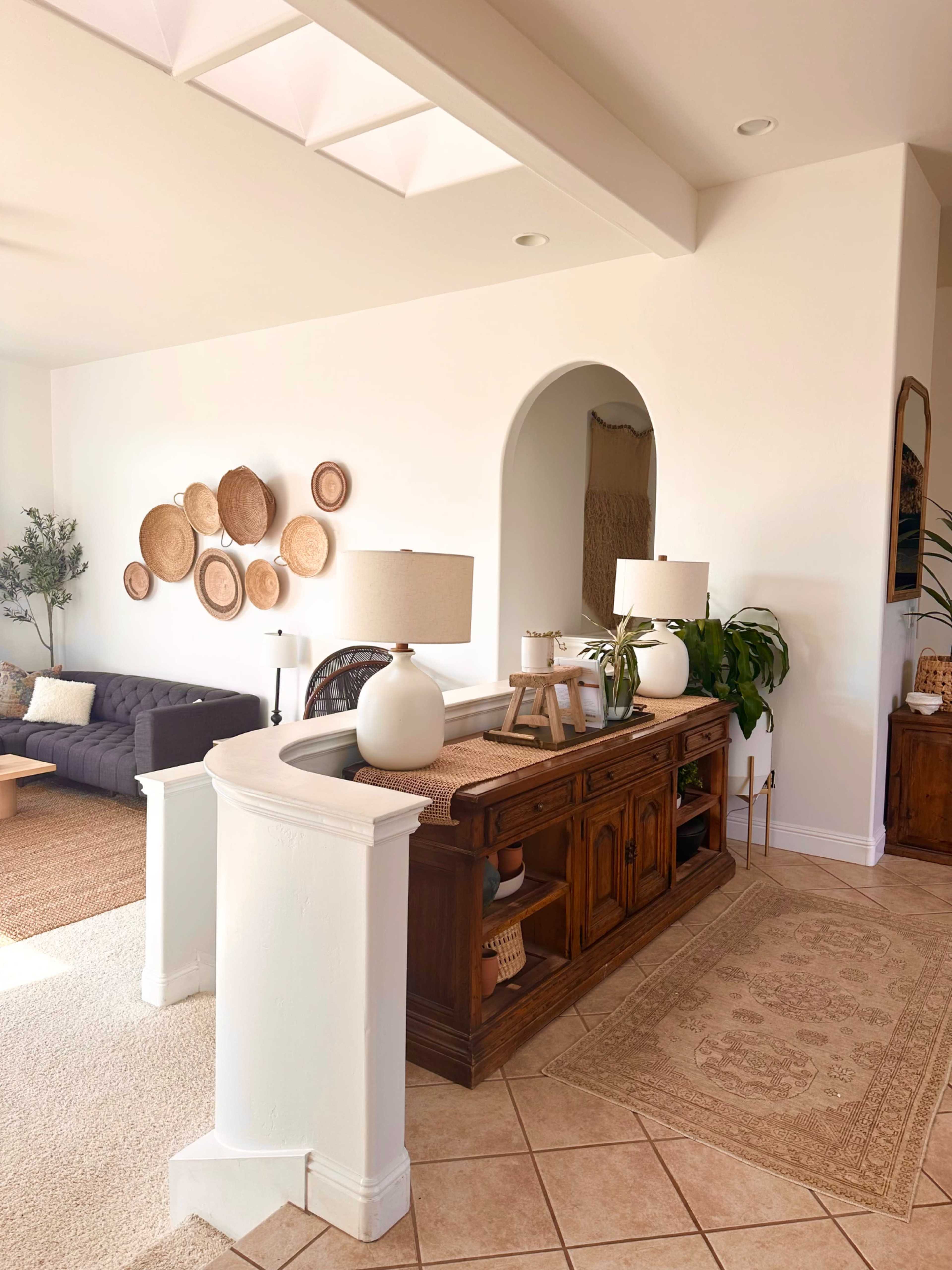 The image shows a bright living space featuring a wooden console table with lamps, decorative wall plates, a dark sofa, and a large area rug.