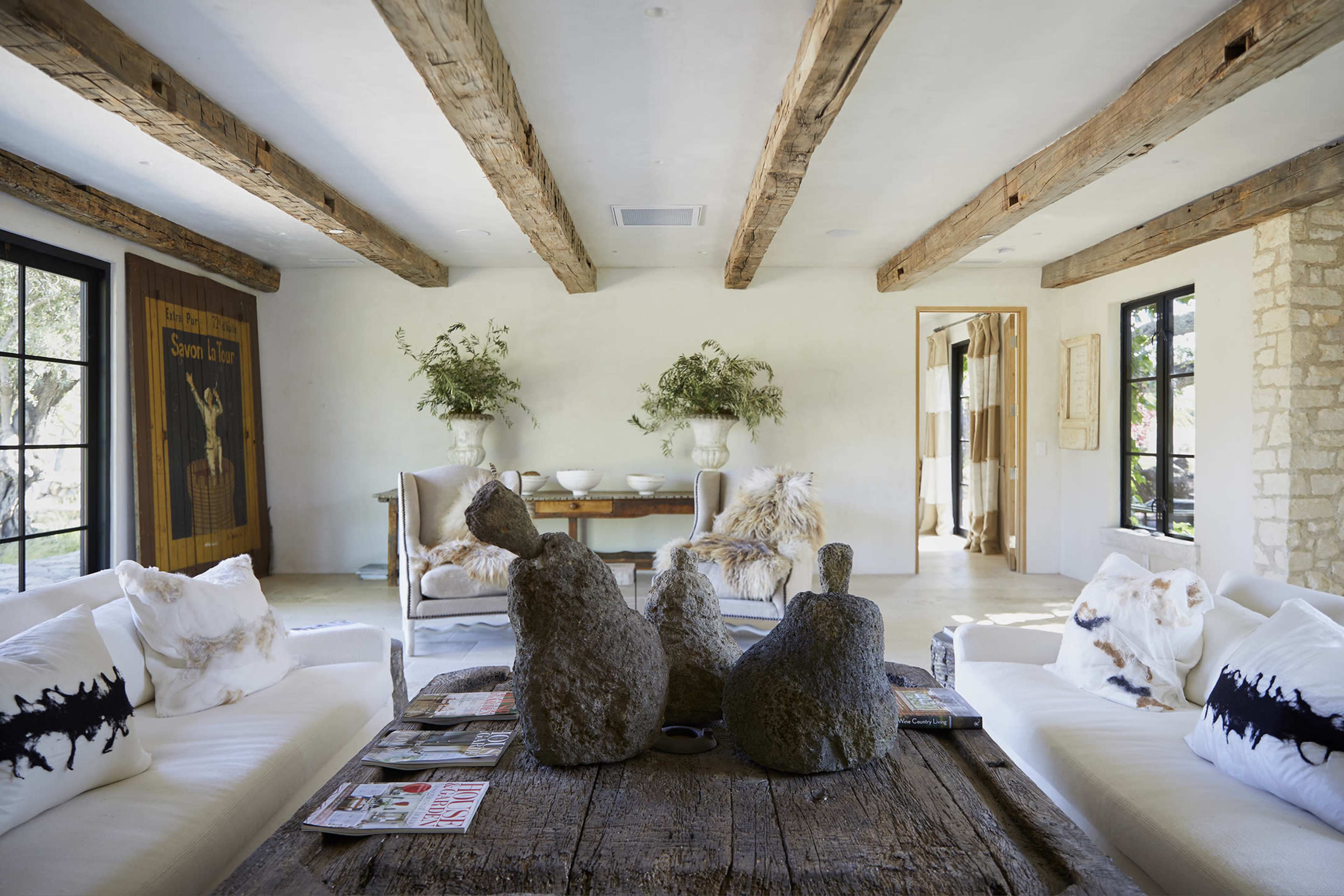 A modern living room with exposed wooden beams, a large rustic table, two textured sculptures, and light-colored furniture arranged around the space.