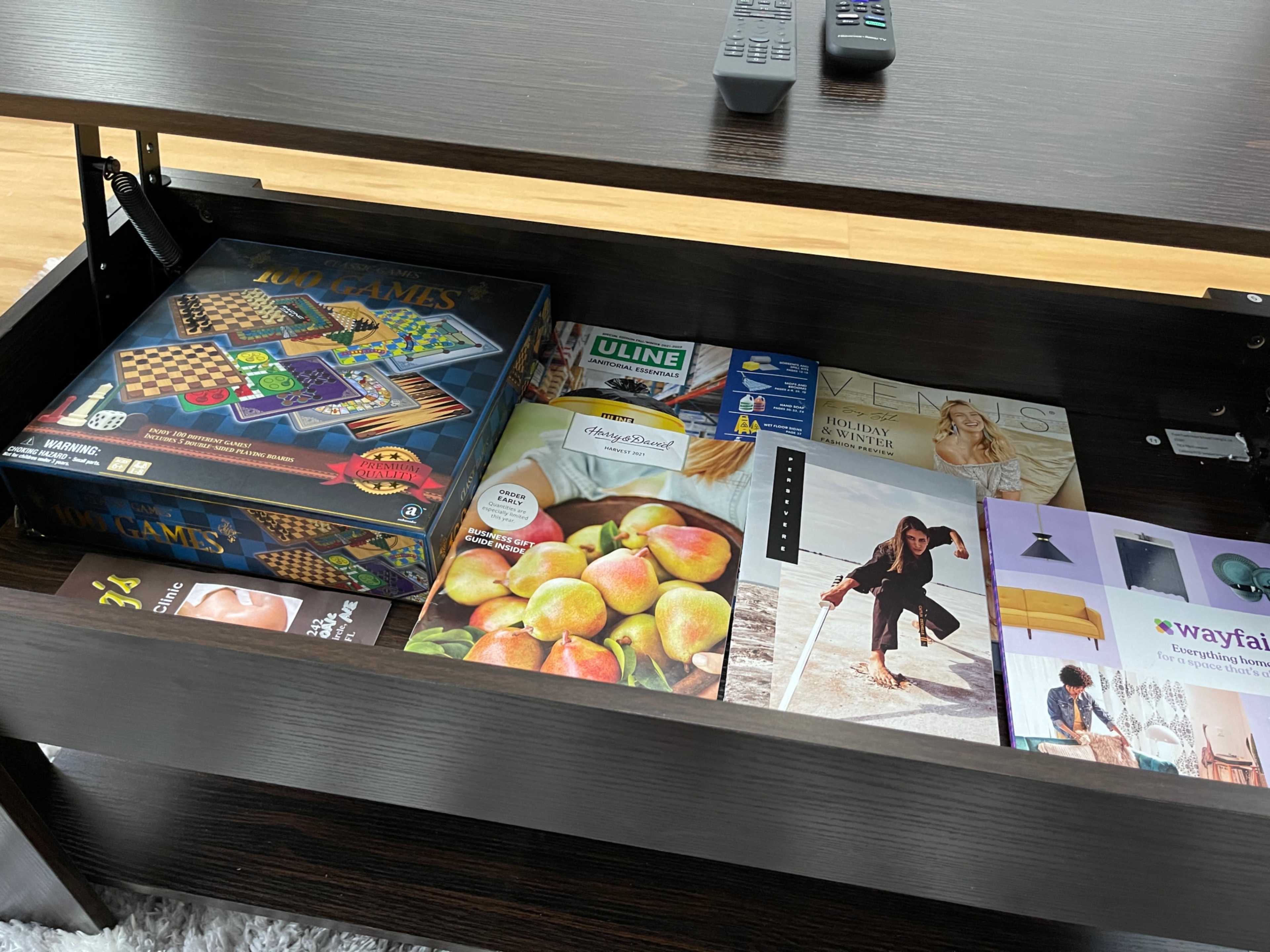 An open coffee table drawer containing a board game, magazines, a box of light bulbs, and a decorative item.