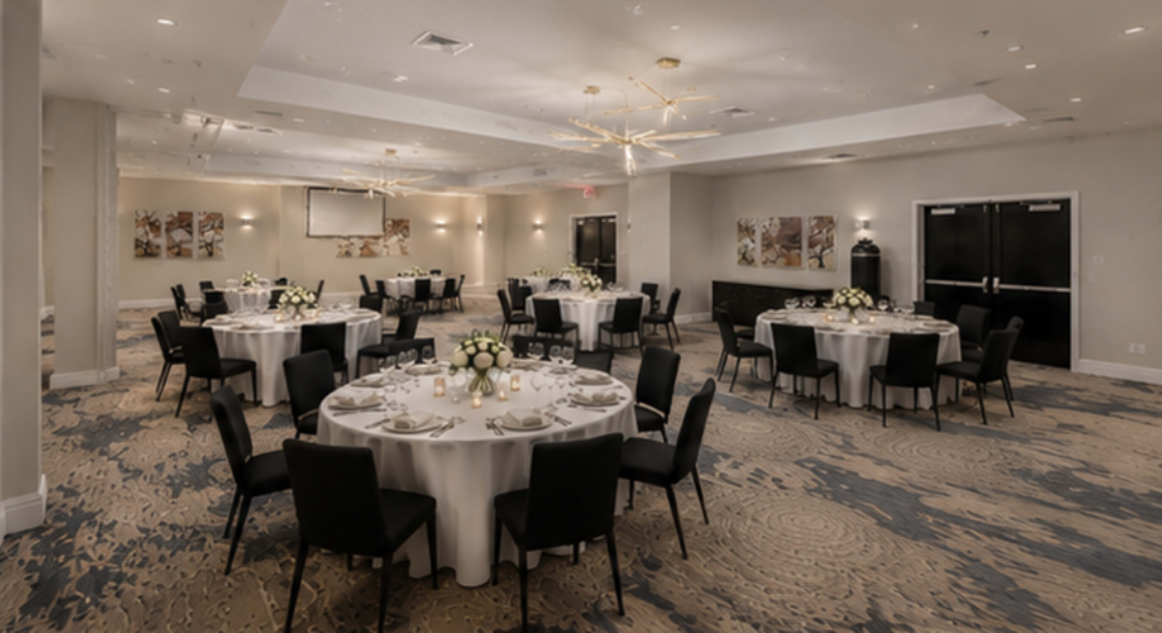 The image shows a spacious event room with several round tables set for dining, decorated with white tablecloths and centerpieces, under modern lighting fixtures.