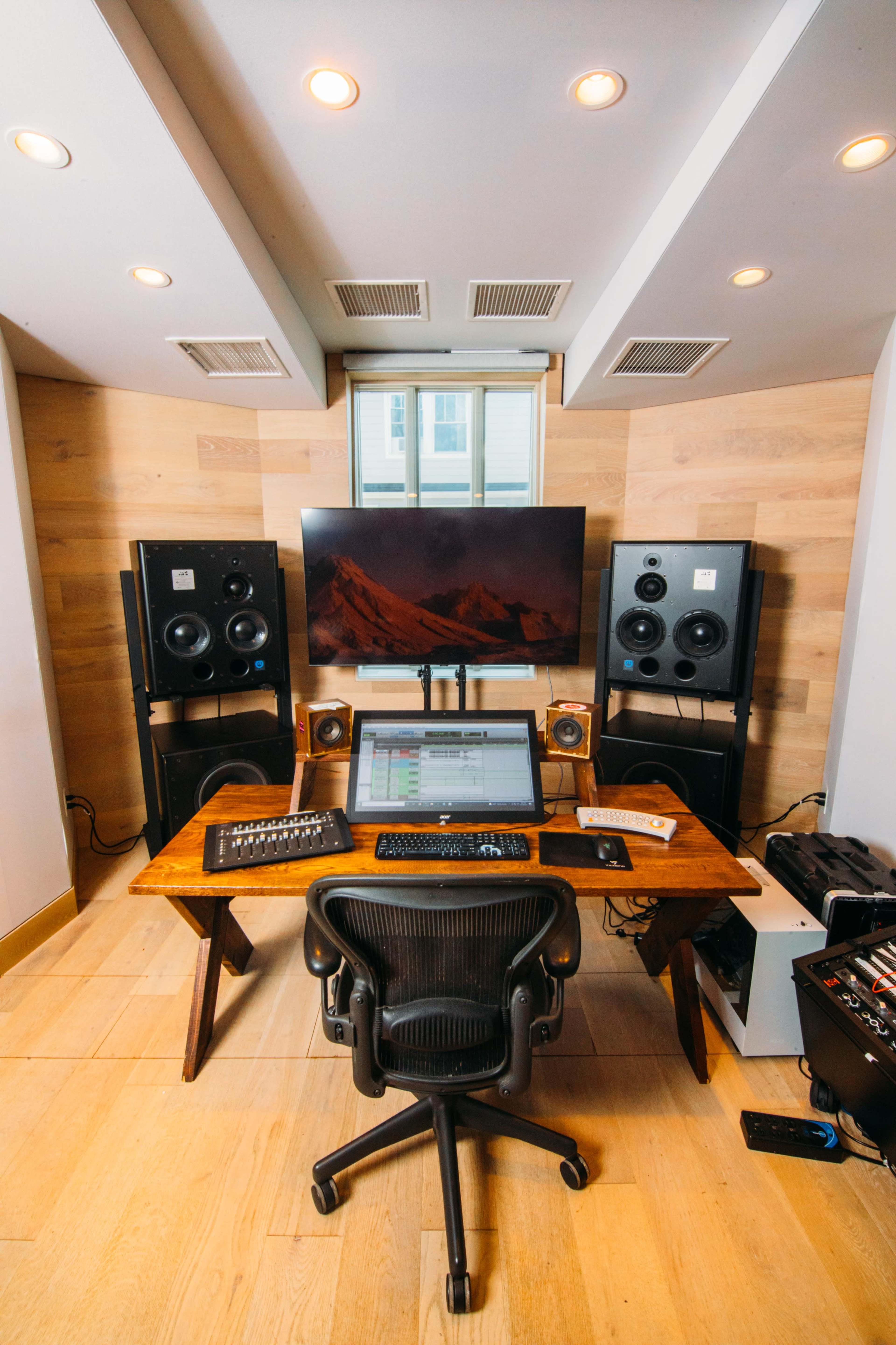 A music production studio featuring a wooden desk with a computer monitor, audio equipment, and speakers, surrounded by light-colored wooden walls.