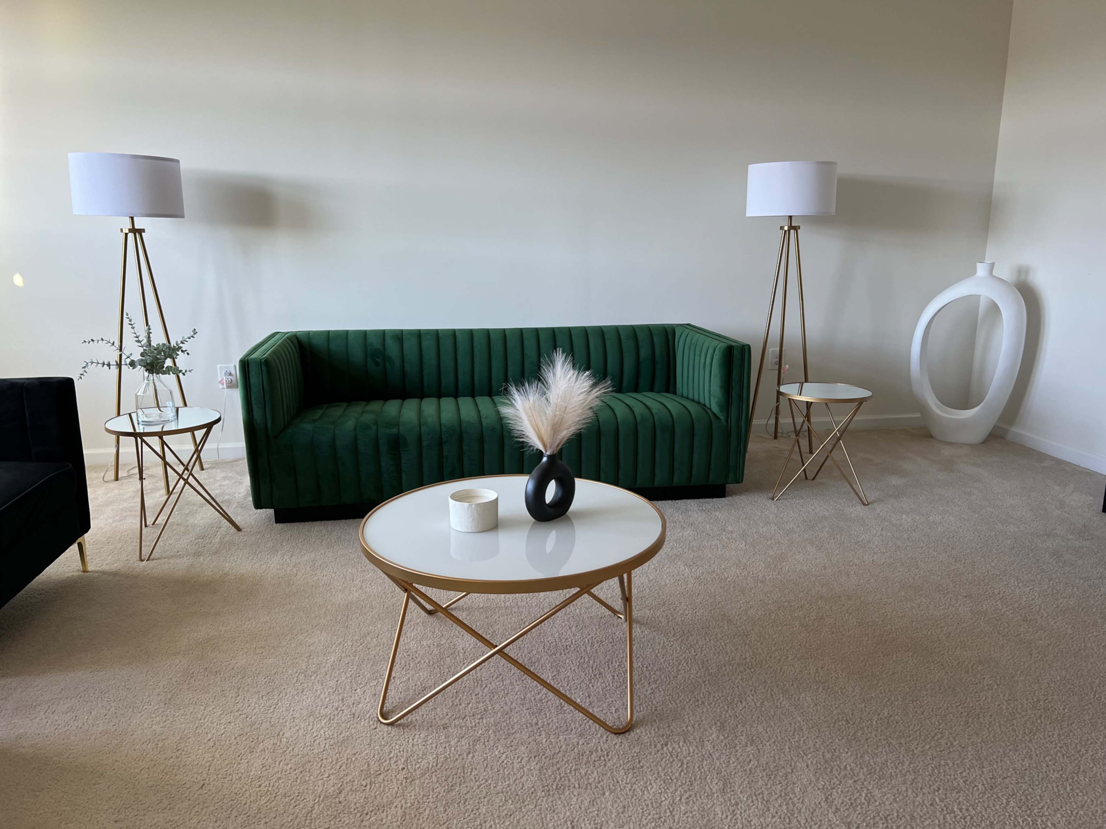 A modern living room featuring a green velvet couch, two minimalist tables with gold bases, a black vase with decorative pampas grass, and floor lamps beside the couch.