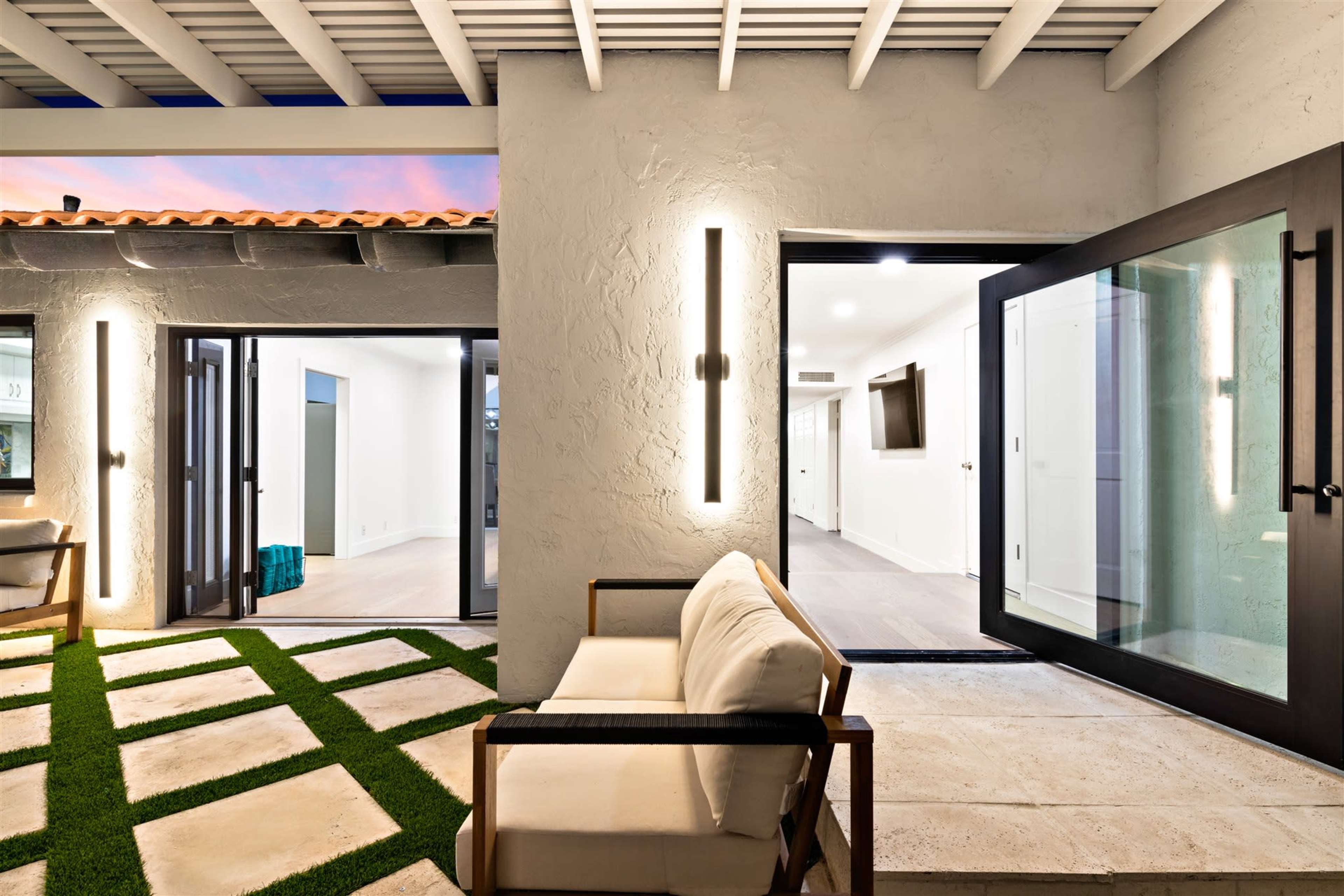 The image shows a modern outdoor seating area with a stone patio, green grass accents, and large glass doors leading to an interior space.