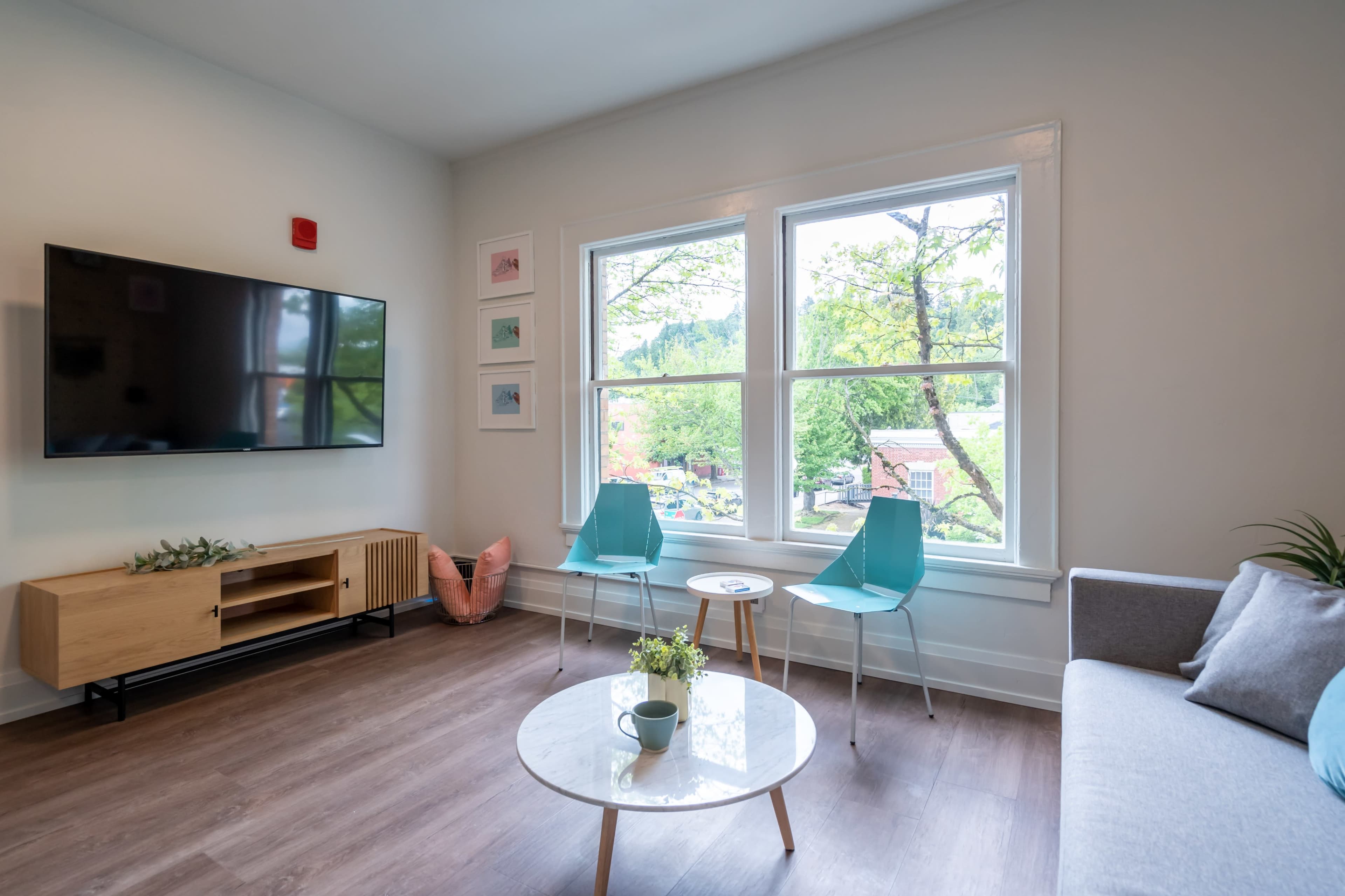 A modern living room features a television mounted on the wall, two turquoise chairs, a round coffee table, and a large window with a view of greenery outside.